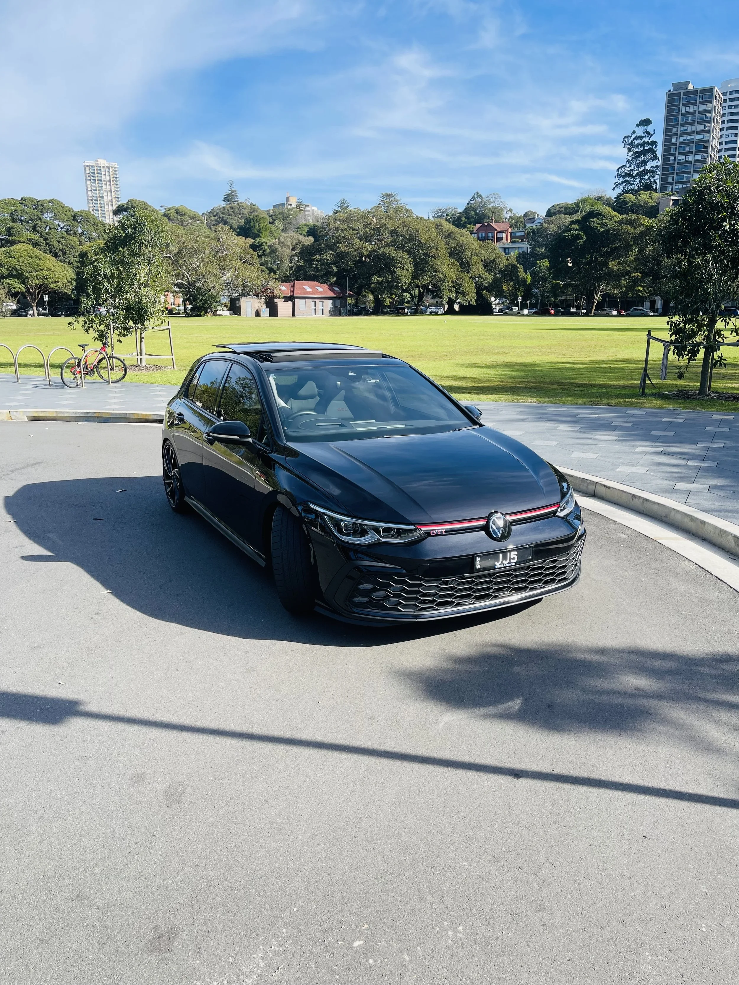 A black Volkswagen car parked on a paved area near green park grass and trees, with tall buildings in the background under a partly cloudy sky.