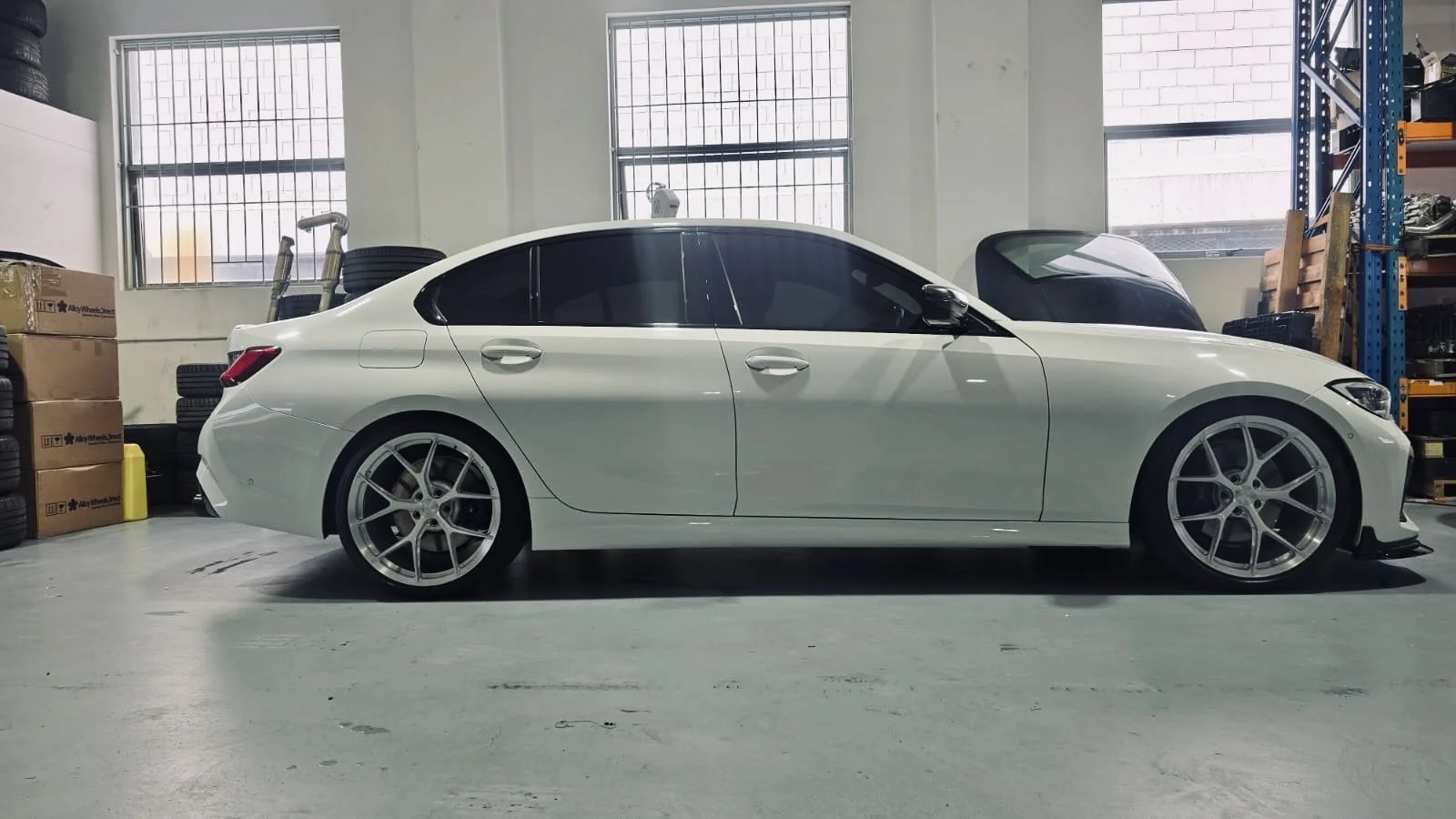 A white luxury sedan car parked inside a garage with windows, tires, and car parts nearby.