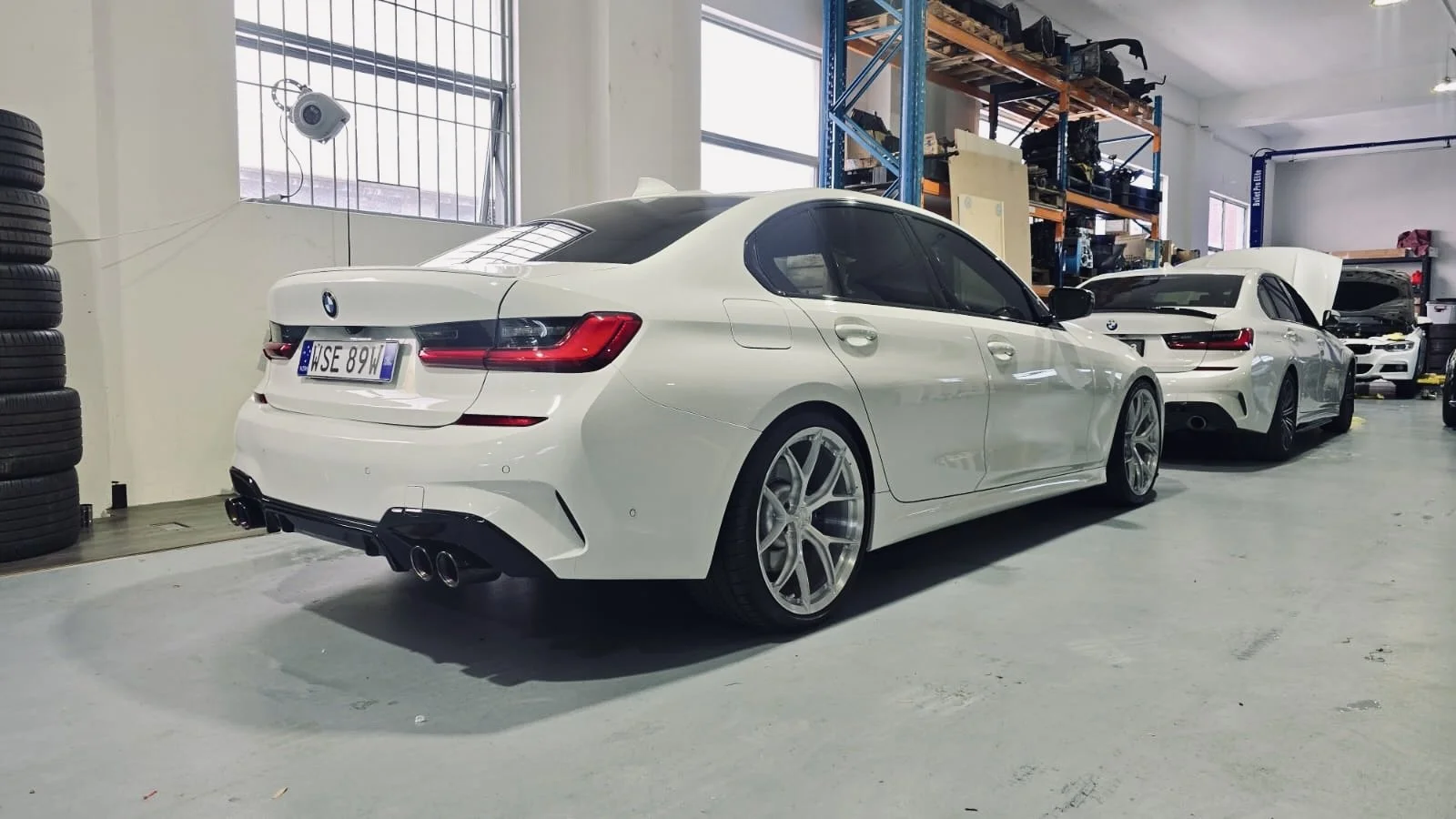 White BMW inside a garage, with multiple luxury cars and automotive parts on shelves.
