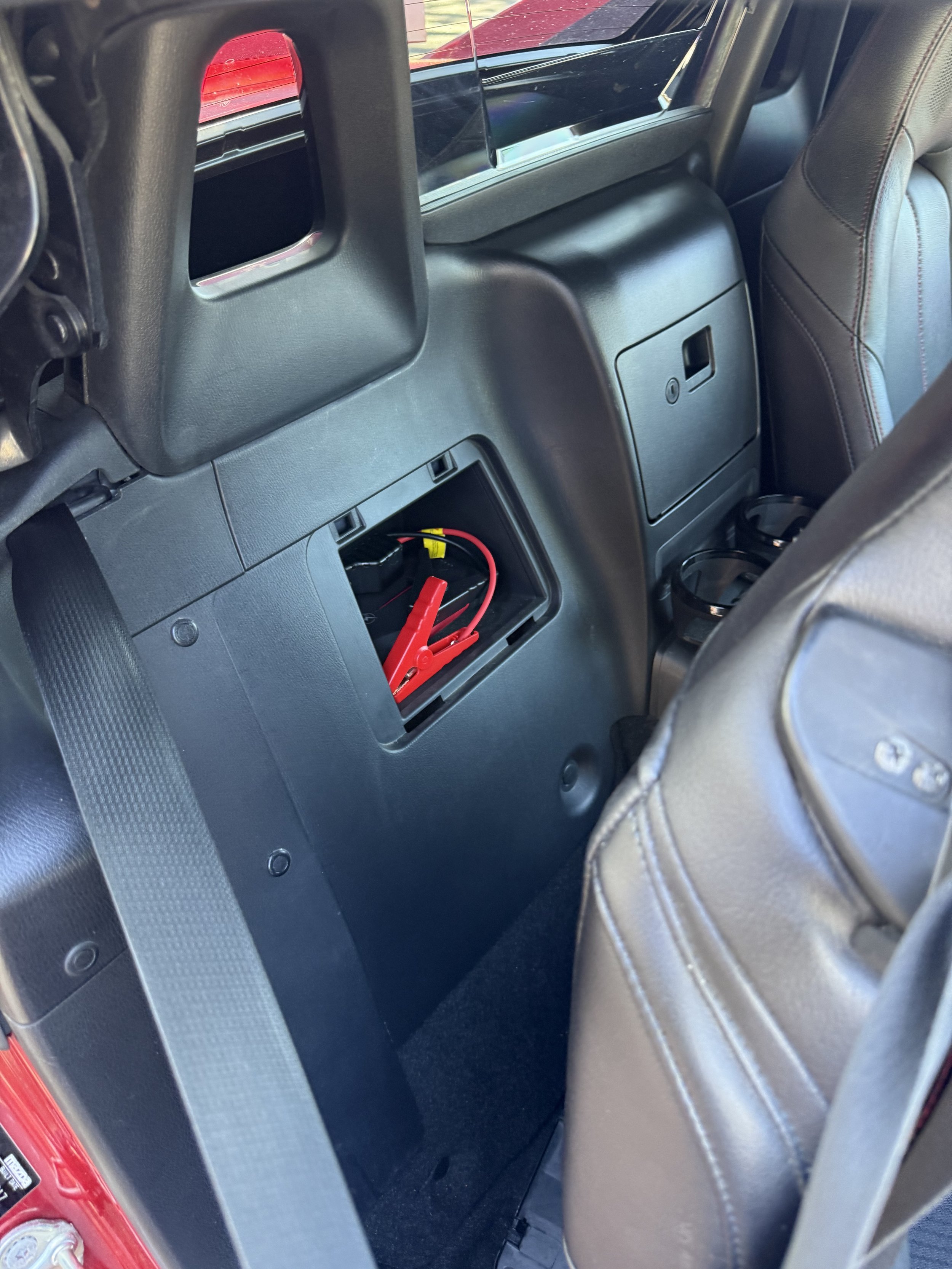 The trunk of a car with a black carpeted surface, showing a small storage compartment with a warning label and a black netted organizer, and part of the car's internal structure.