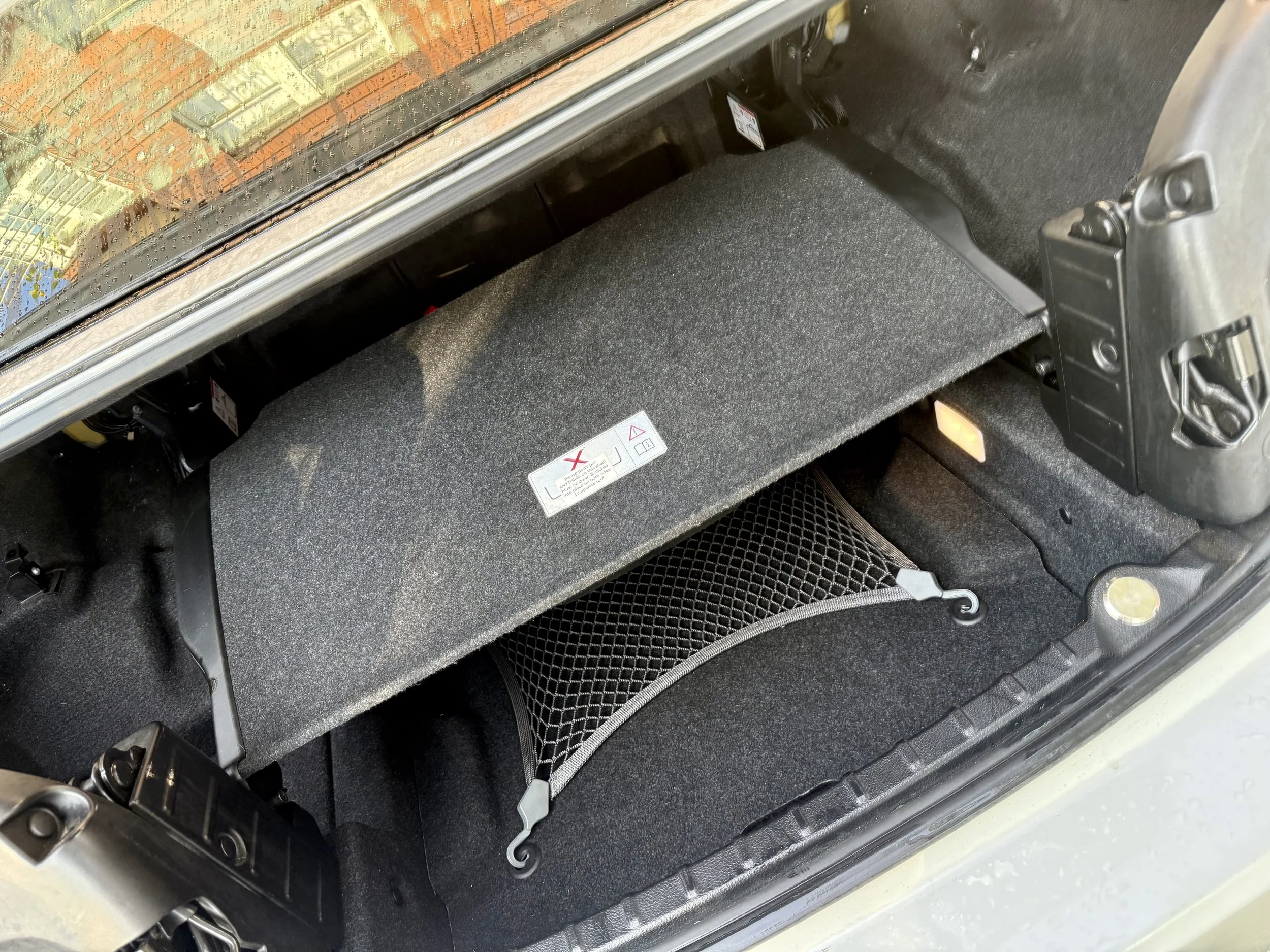 The trunk of a car with a black carpeted surface, showing a small storage compartment with a warning label and a black netted organizer, and part of the car's internal structure.