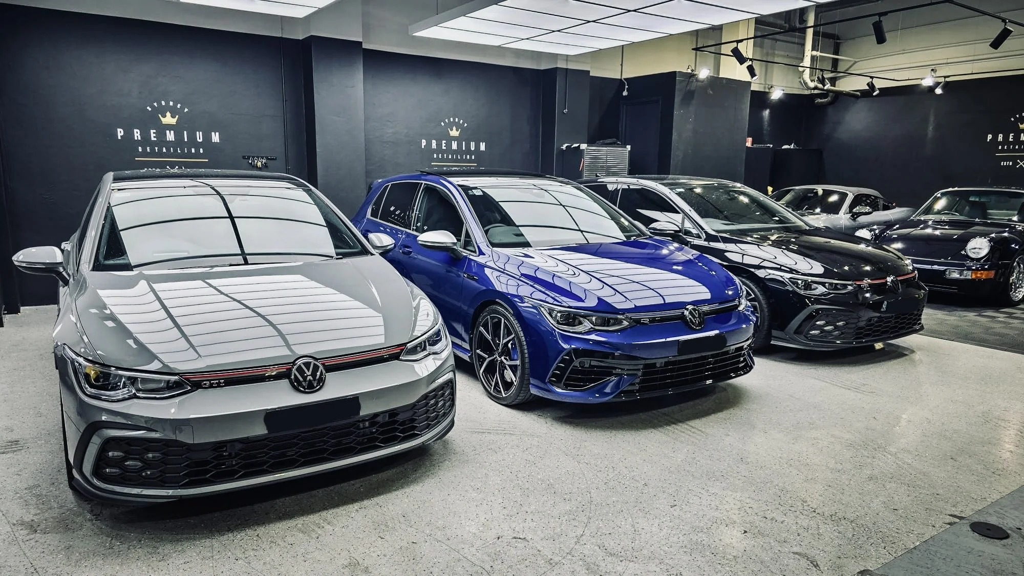A row of luxury cars on display inside a dealership, including a silver Volkswagen Golf GTI, a blue Volkswagen Golf R, and a black Porsche 911, with dark walls and branding for Premium Detailing.