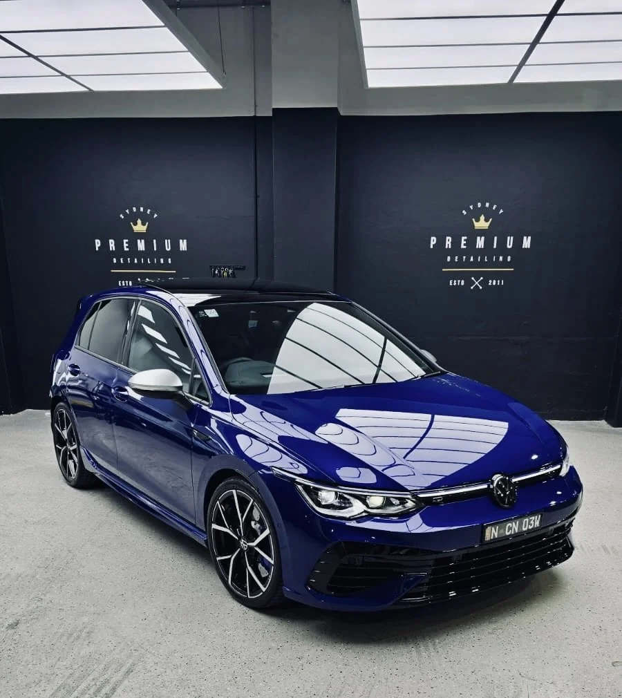 A blue Volkswagen car with black rims inside a showroom with black walls and ceiling panels, featuring the words 'Sydney Premium Detailing' and an image of a crown on the walls.