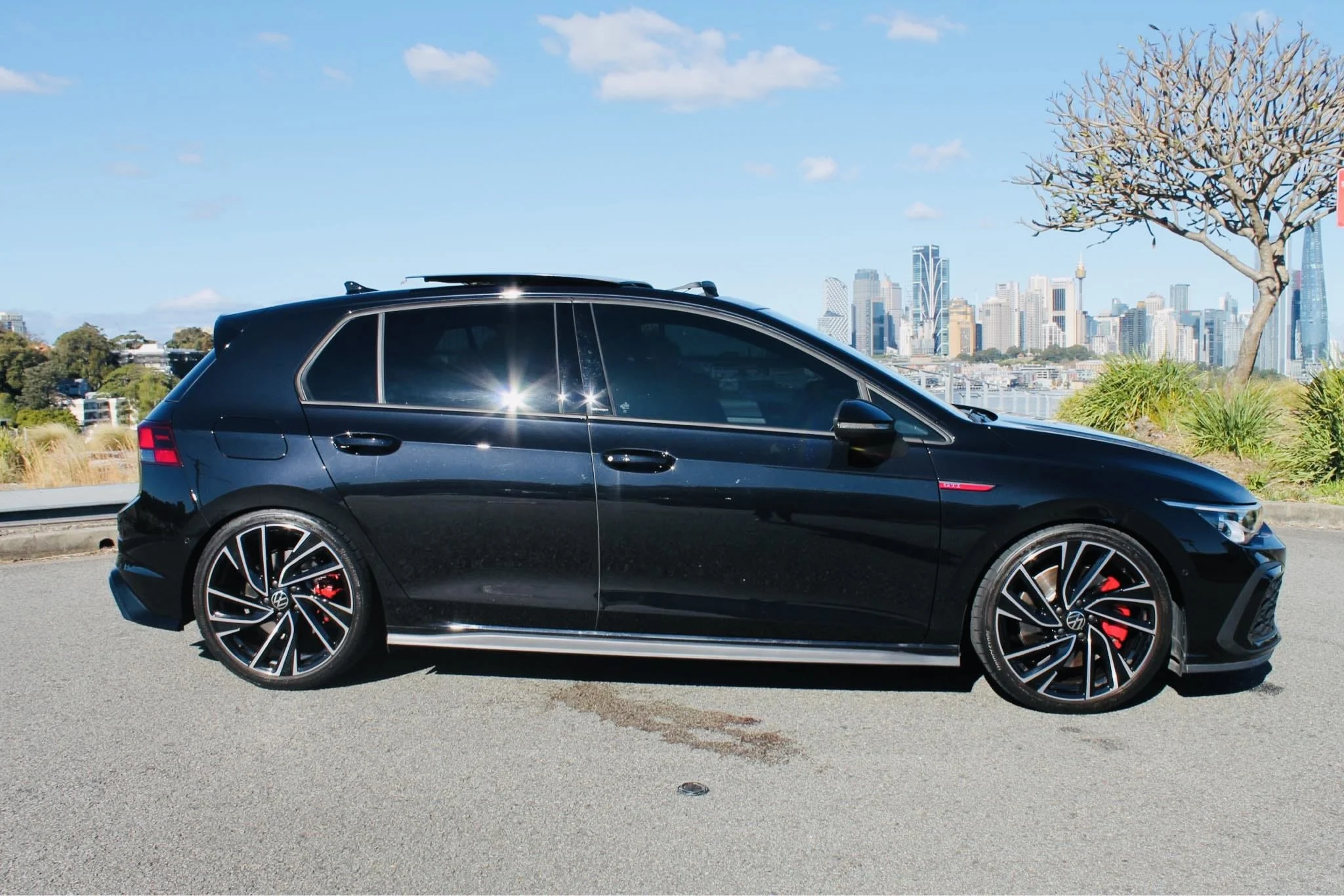 Black Volkswagen hatchback car parked outdoors with a city skyline in the background, clear blue sky, and a tree to the right.