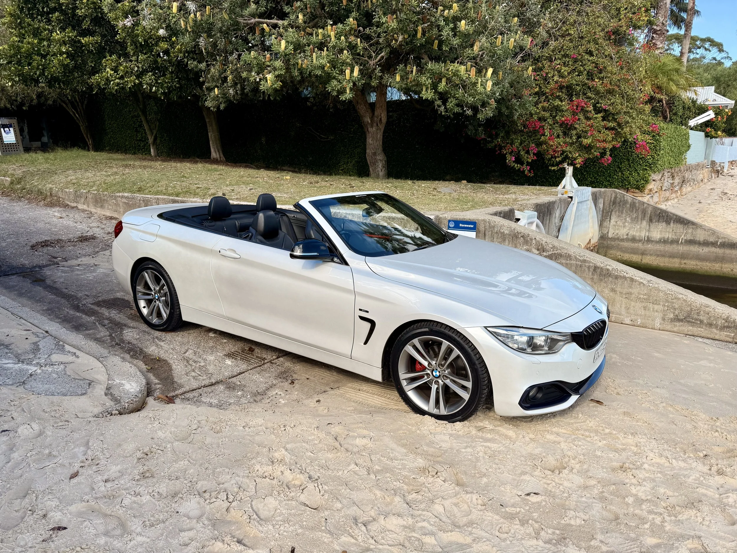A white BMW convertible car parked on a sandy area near the water, with green trees and pink flowers in the background.