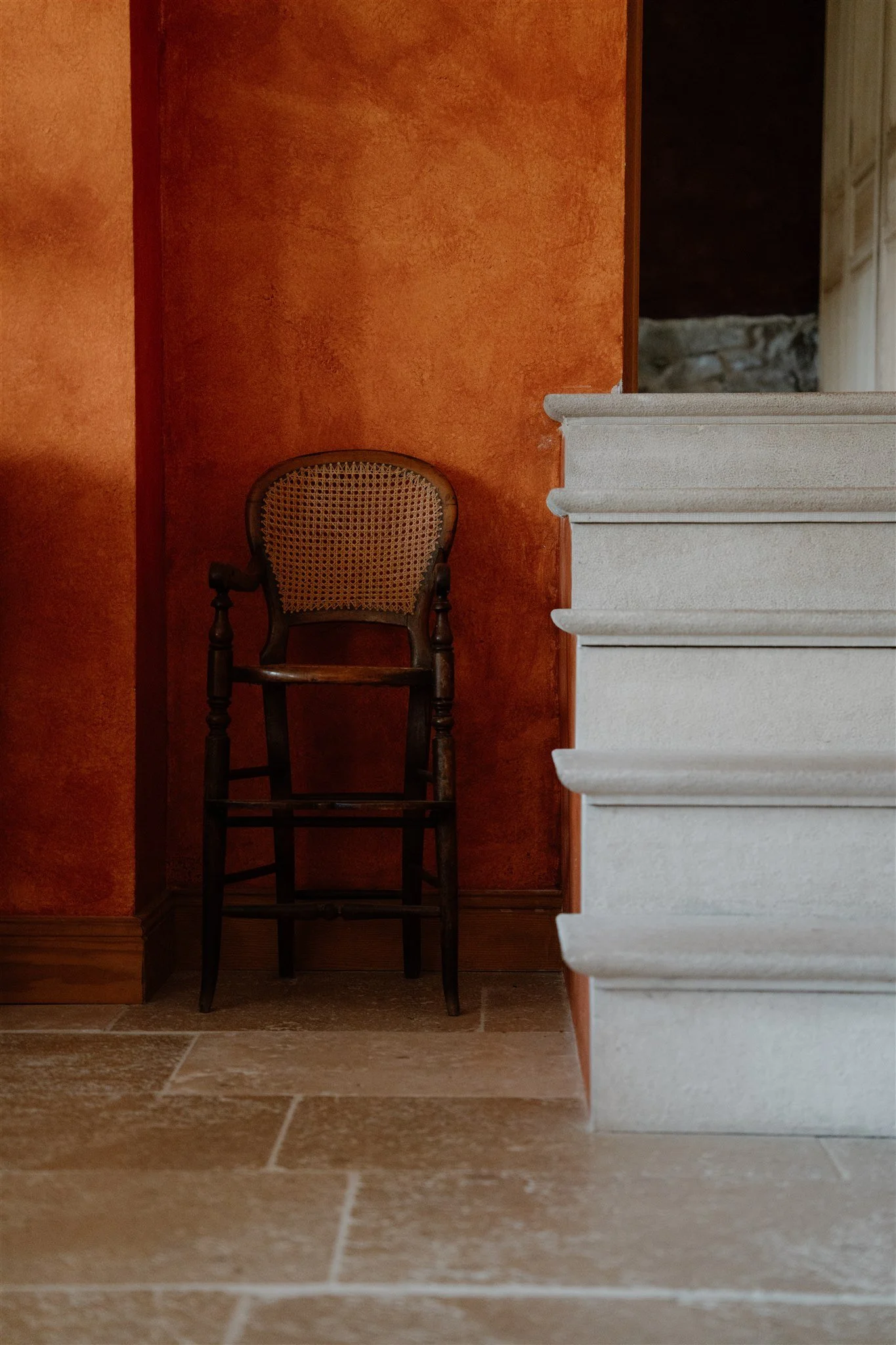 Interior scene with a dark wooden chair featuring a woven cane backrest, a textured orange wall, and a staircase with white steps on the right side, with a tiled floor.