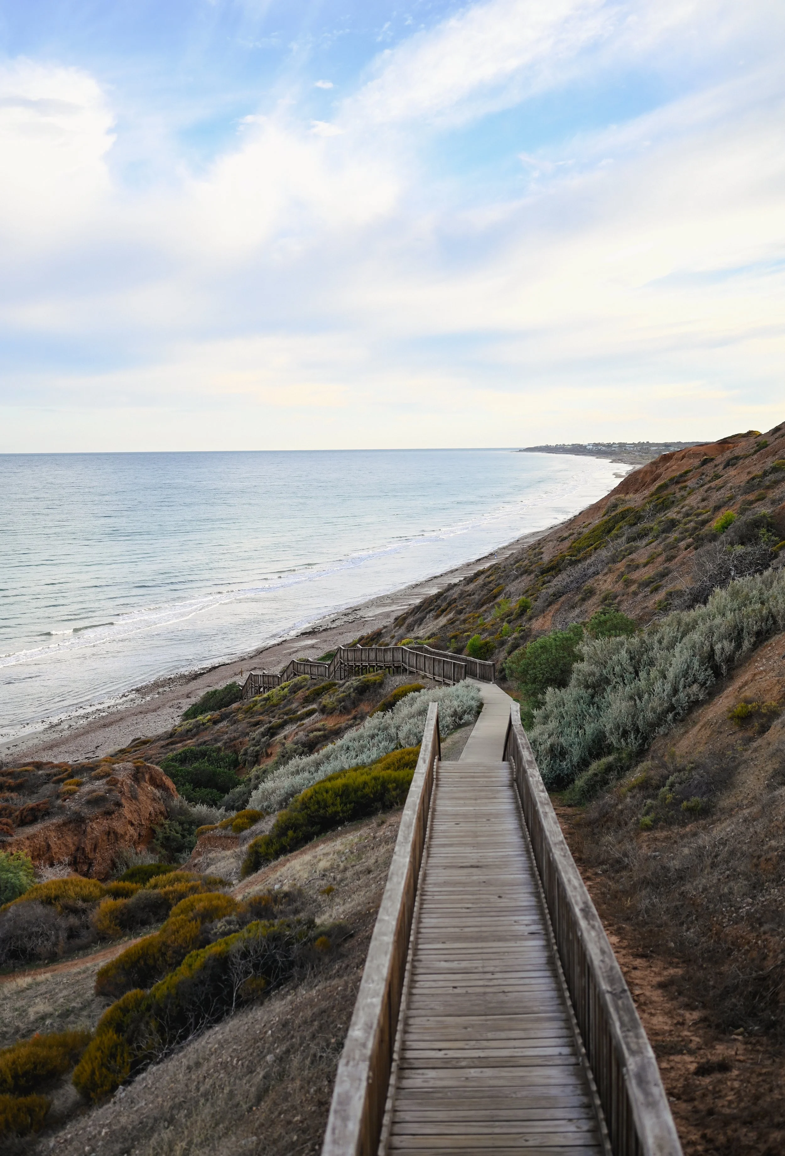 Wooden walkway descending a hillside towards the beach and ocean under a cloudy sky.