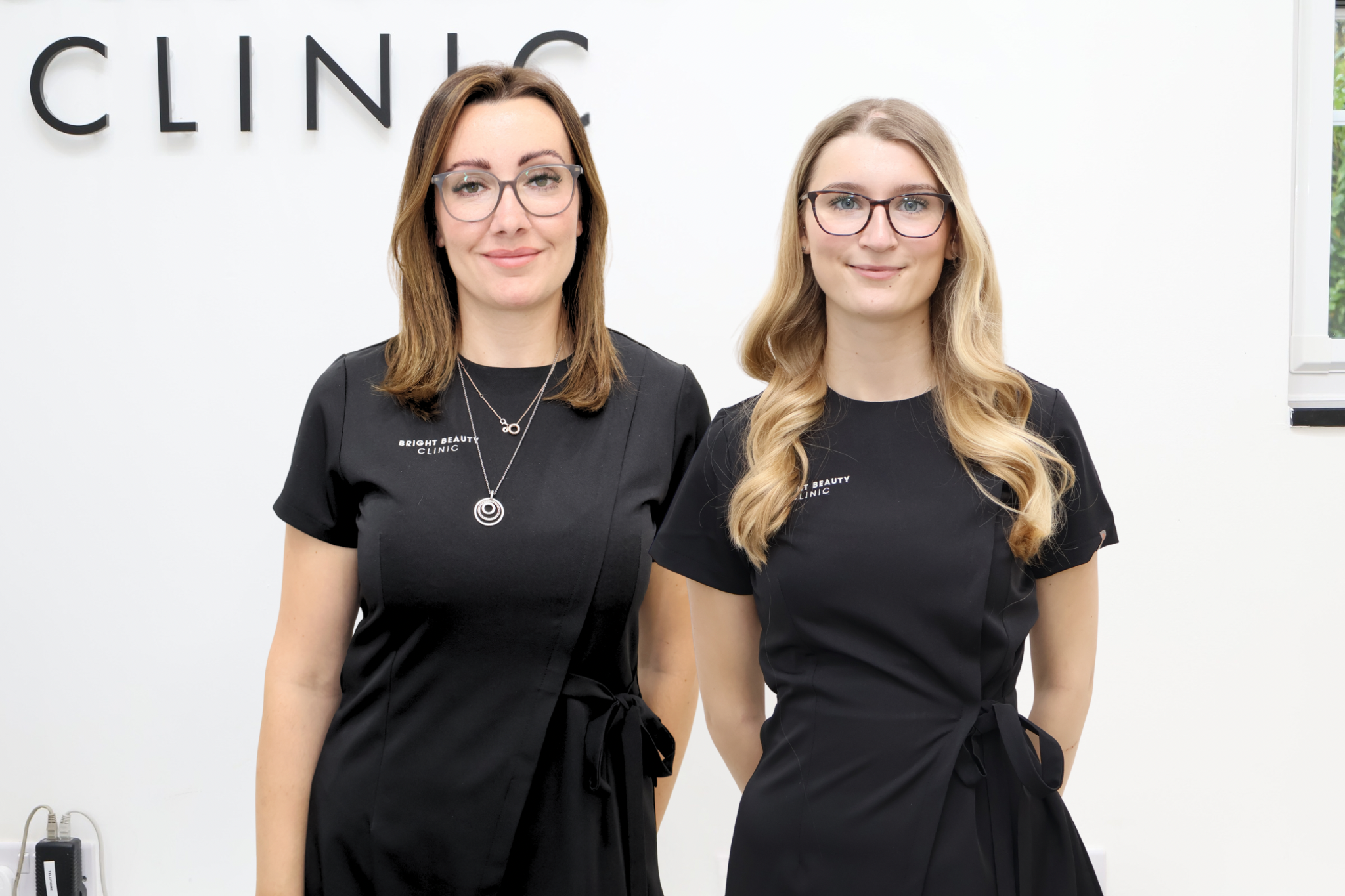 Two women in black scrubs standing in front of a white wall with partial view of the word "CLINIC" visible. Both women are wearing glasses and have long hair, smiling at the camera.