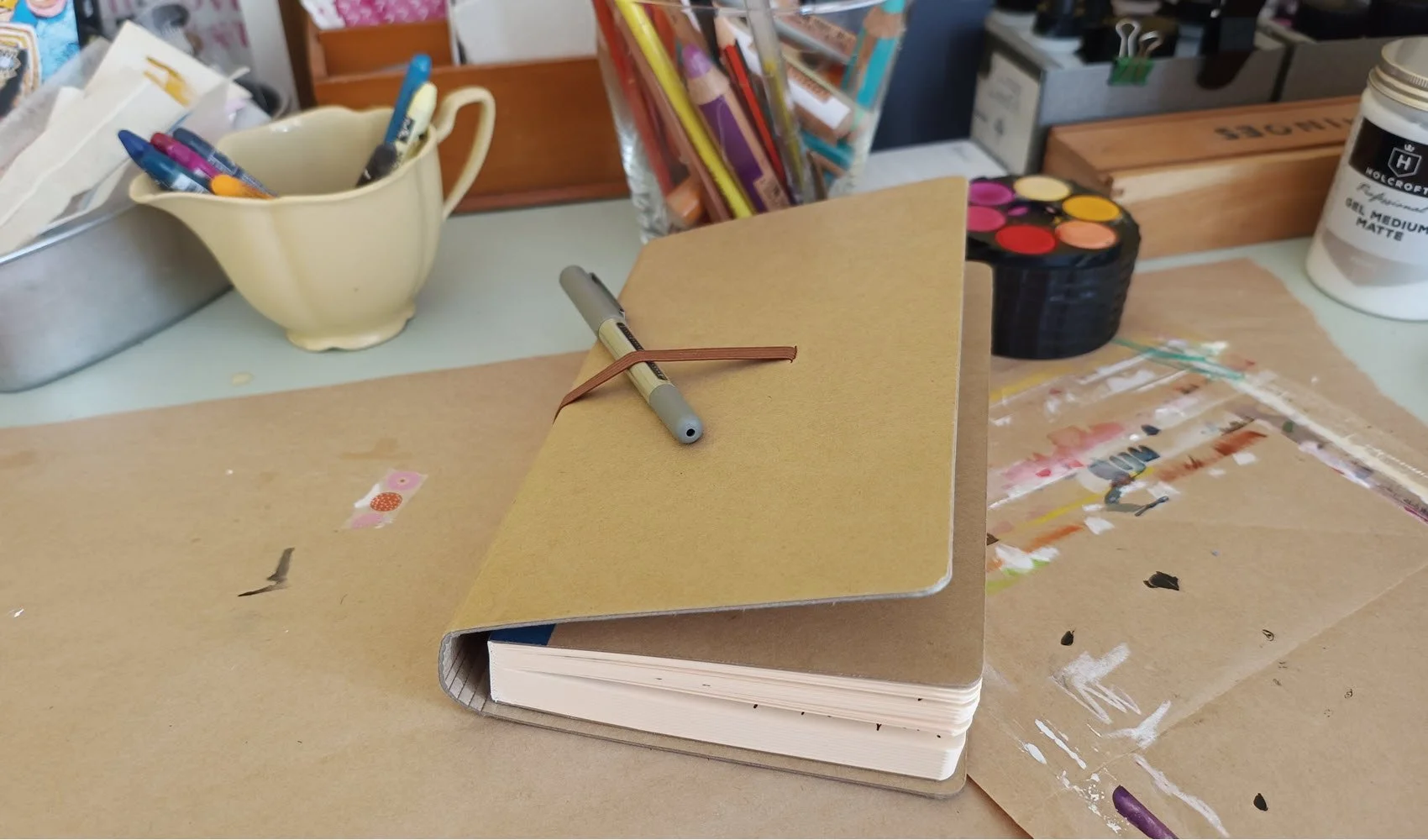 a kraft coloured journal on a table with art materials in the background