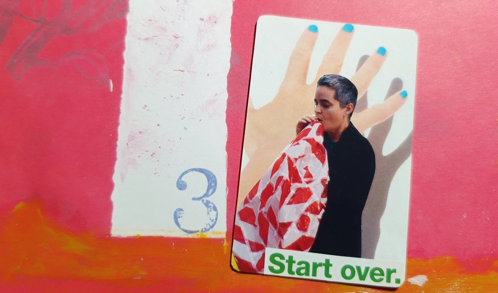A collaged playing card on red background. The collage shows a woman blowing into a balloon and the words start over.