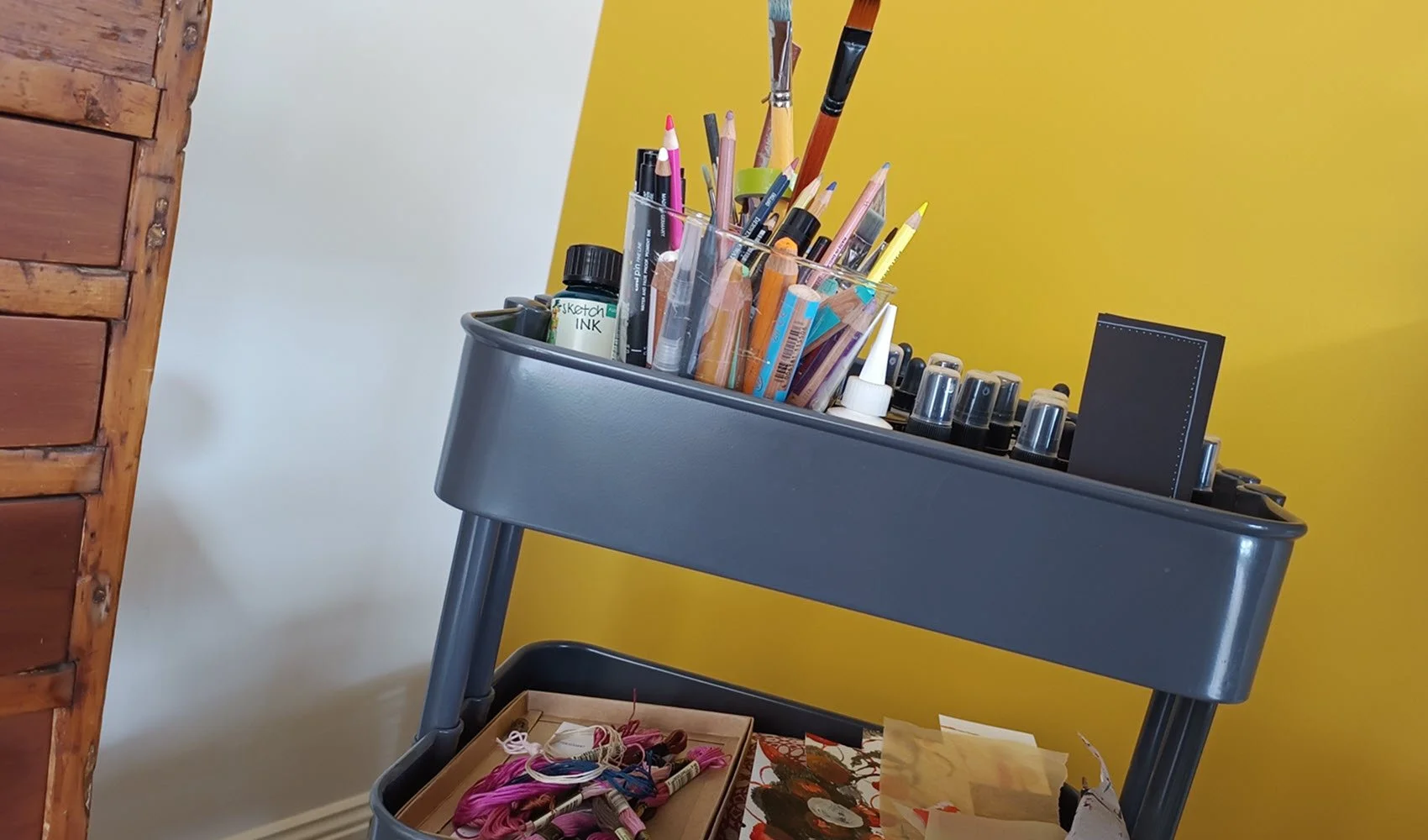 a grey metal trolley filled with art materials like brushes, inks, pencils, glue and yarn in front of a yellow wall