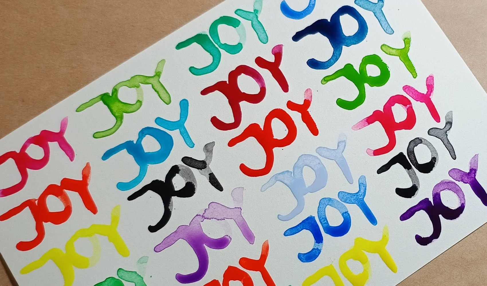 The word JOY written in watercolour in all colours of the rainbow