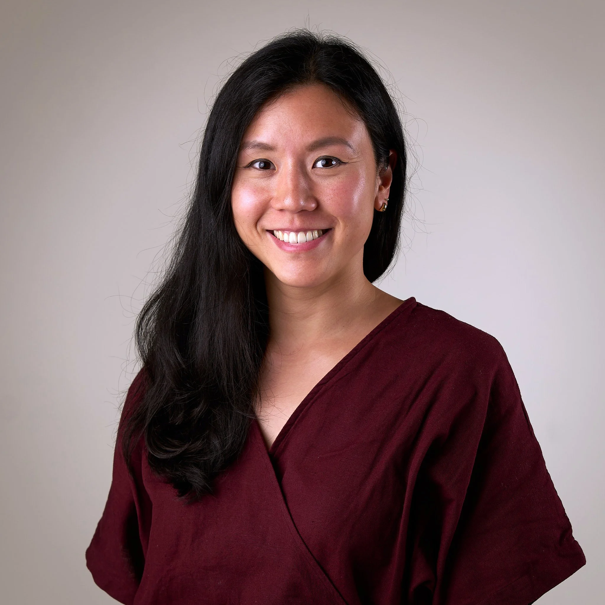 A woman with long dark hair smiling, wearing a maroon top, against a plain gray background.