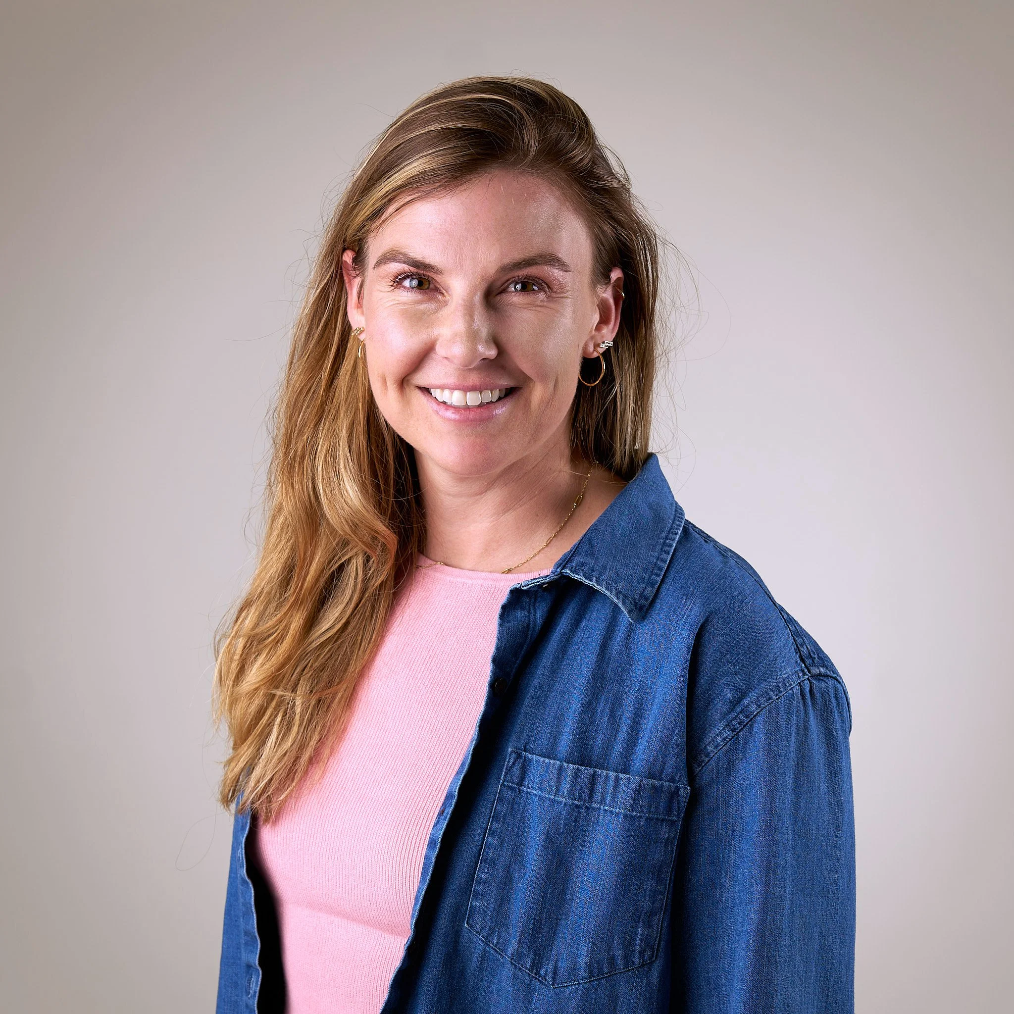 A smiling woman with light brown hair, wearing hoop earrings, a pink shirt, and a blue denim jacket, standing against a neutral background.