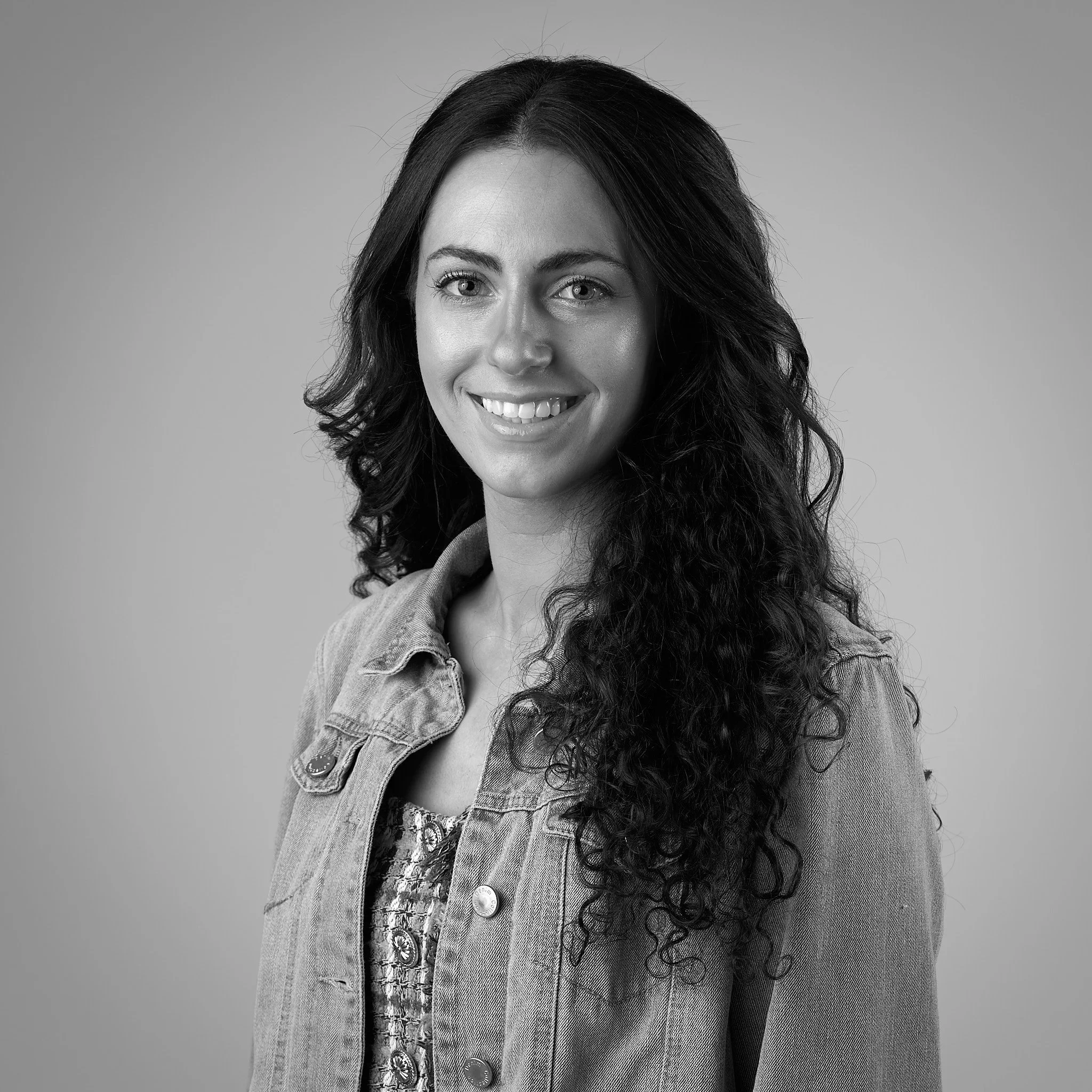 Close-up portrait of a woman with long curly hair, wearing a denim jacket, smiling.