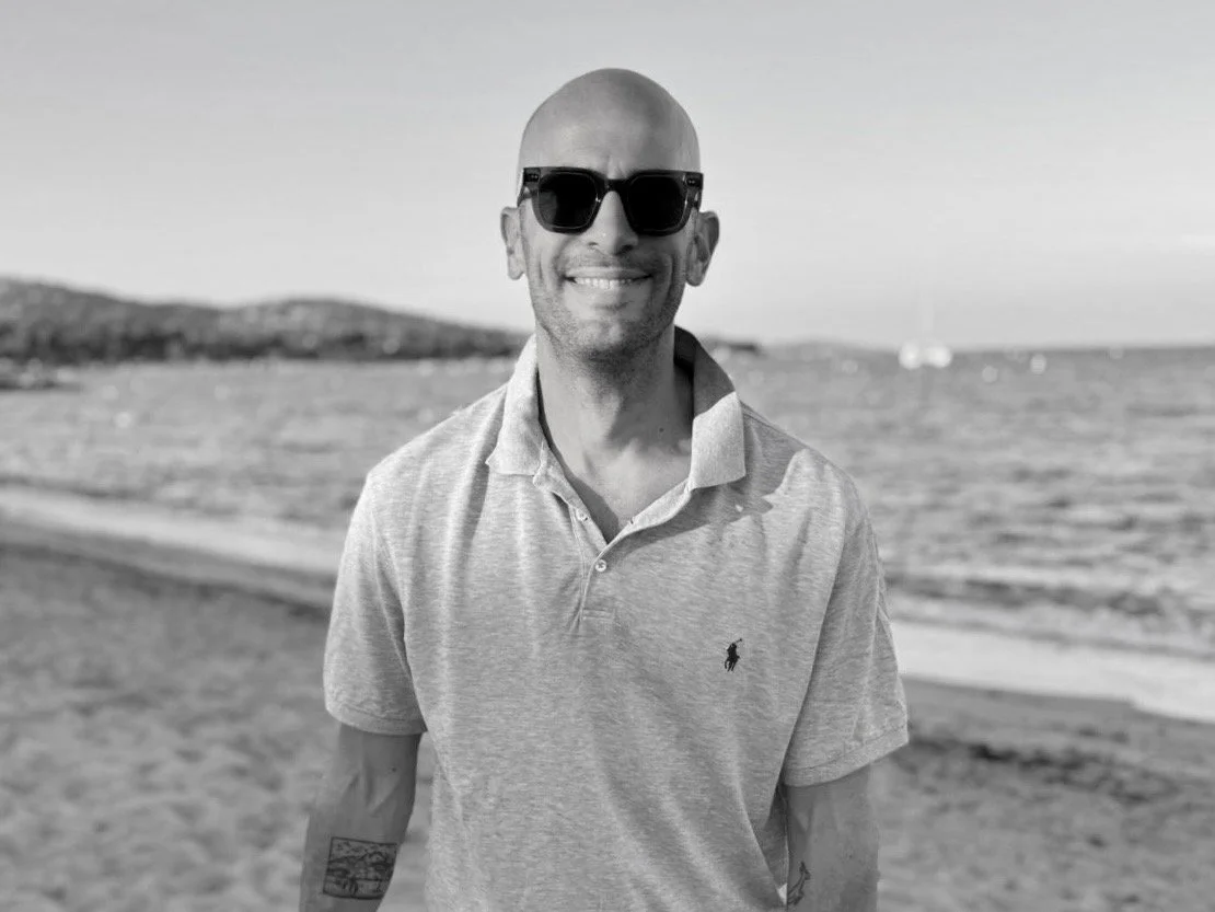 A smiling man wearing sunglasses and a light-colored polo shirt at the beach with the ocean and distant land in the background.