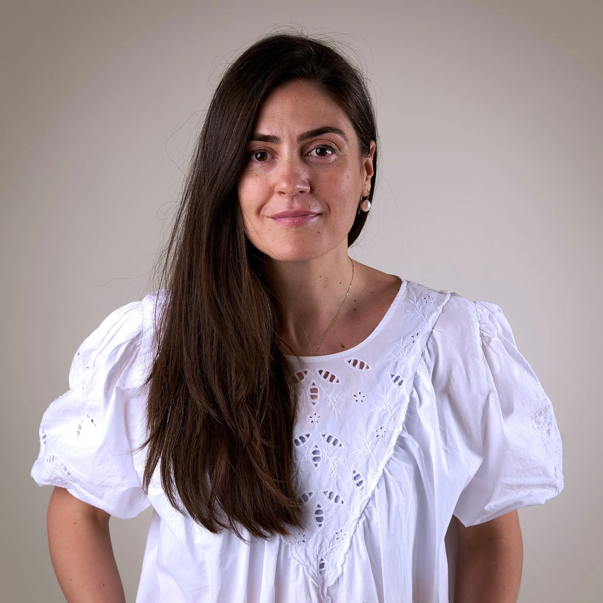 Close-up of a woman with long dark hair wearing a white eyelet blouse and pearl earrings, standing against a plain neutral background.