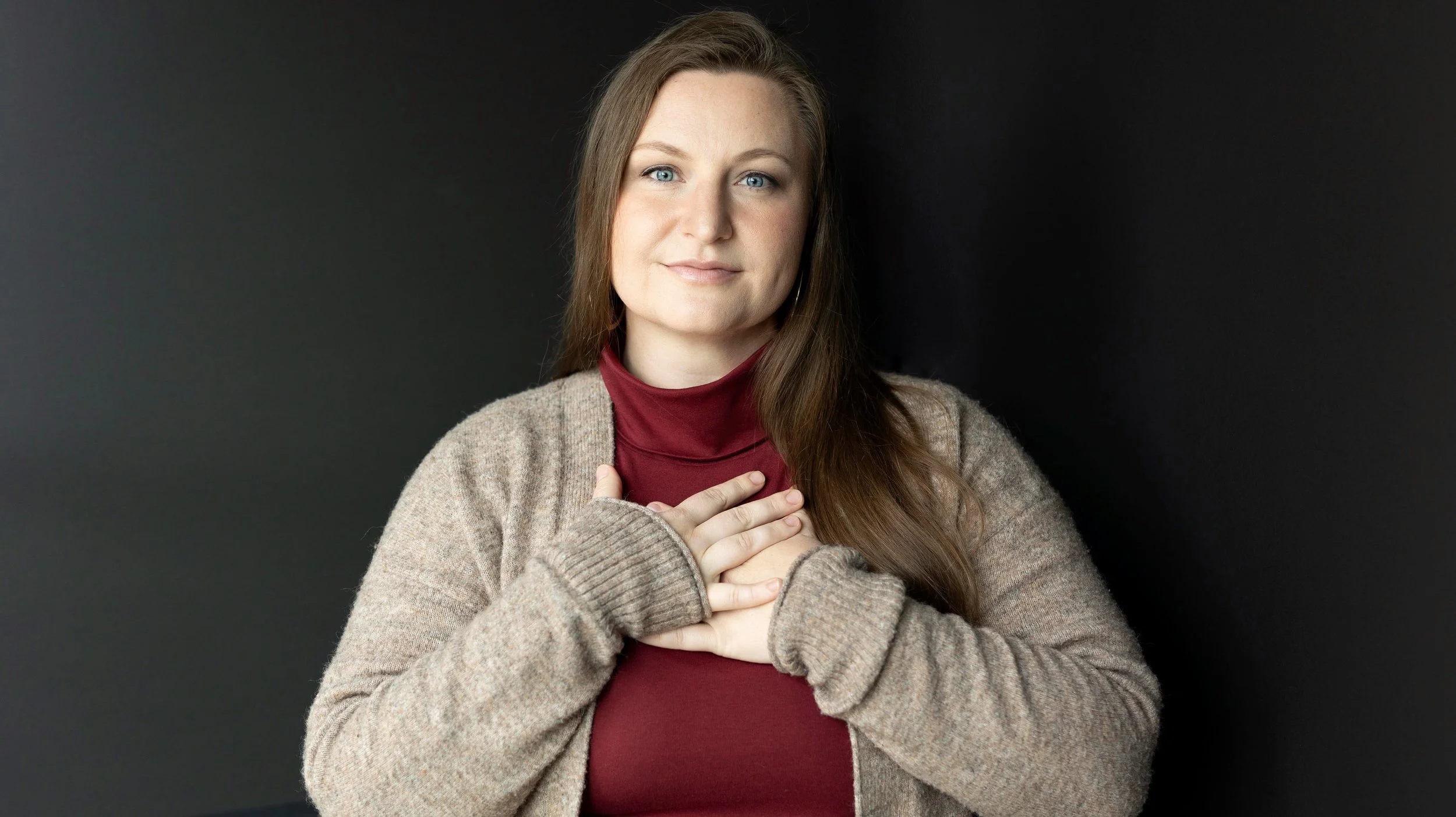A woman with long brown hair and blue eyes holds her hands against her heart, standing against a dark background.