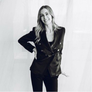 A woman with long wavy hair smiling, wearing a black blazer and black pants, standing against a plain background.