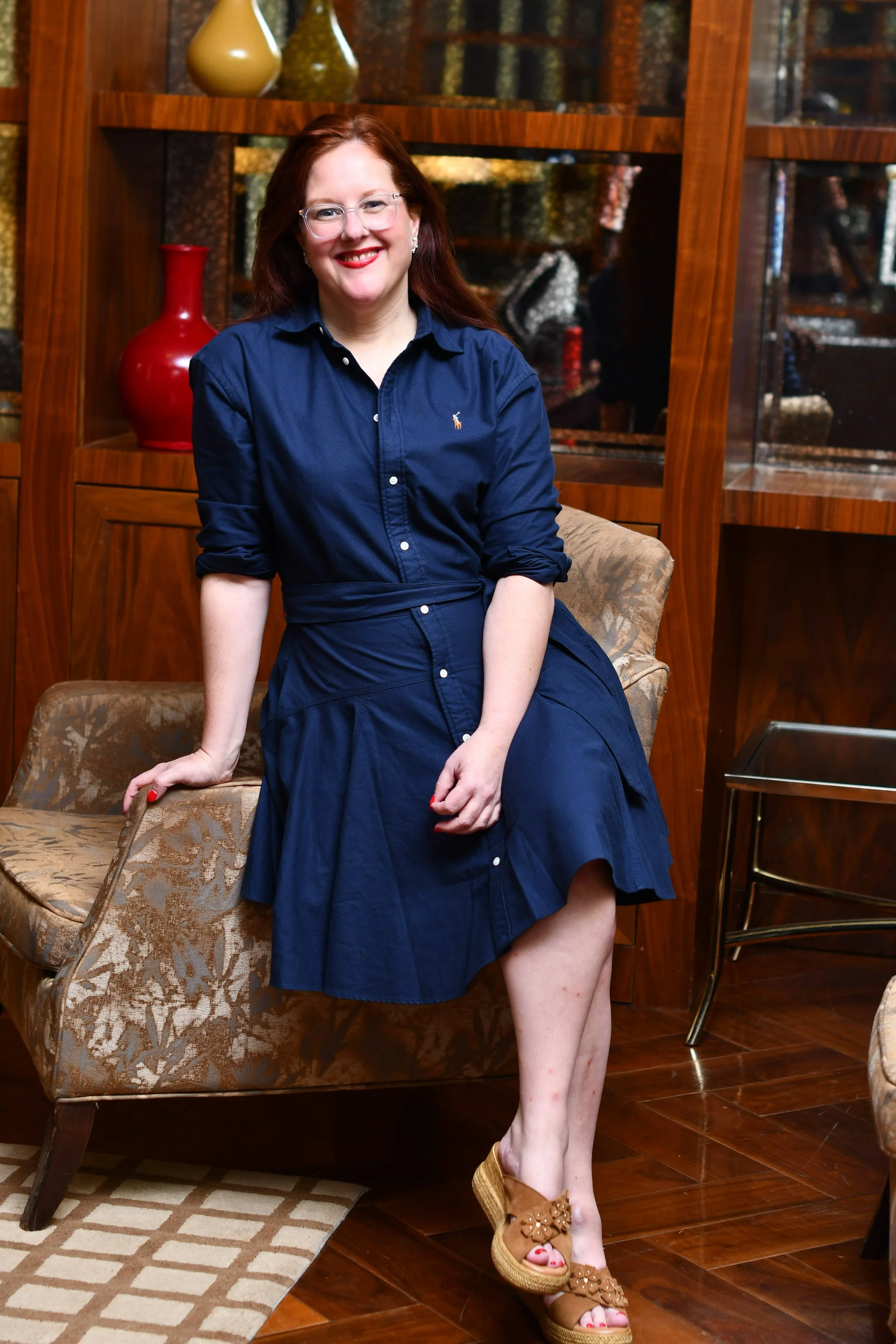 A woman with red hair and glasses smiling outdoors.