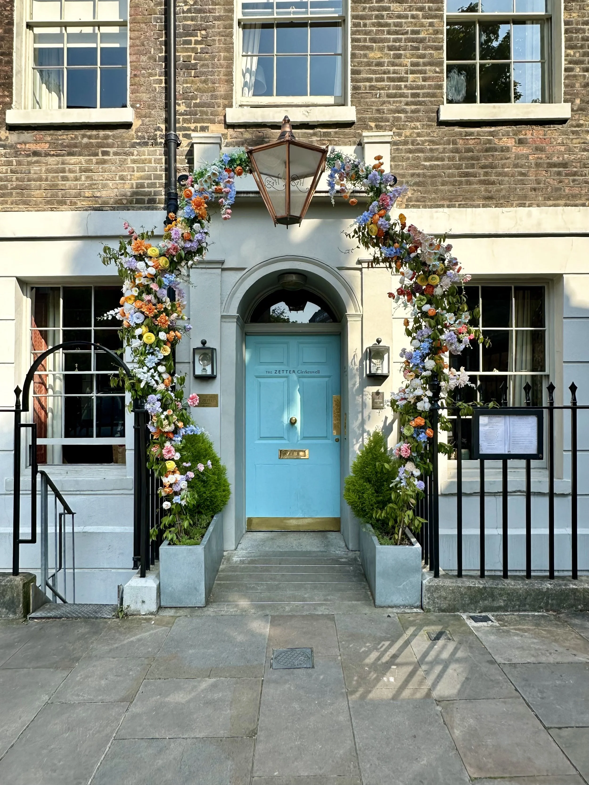 A doorway with a bright blue door, floral arch, and small trees in planters on either side, outside a brick building.