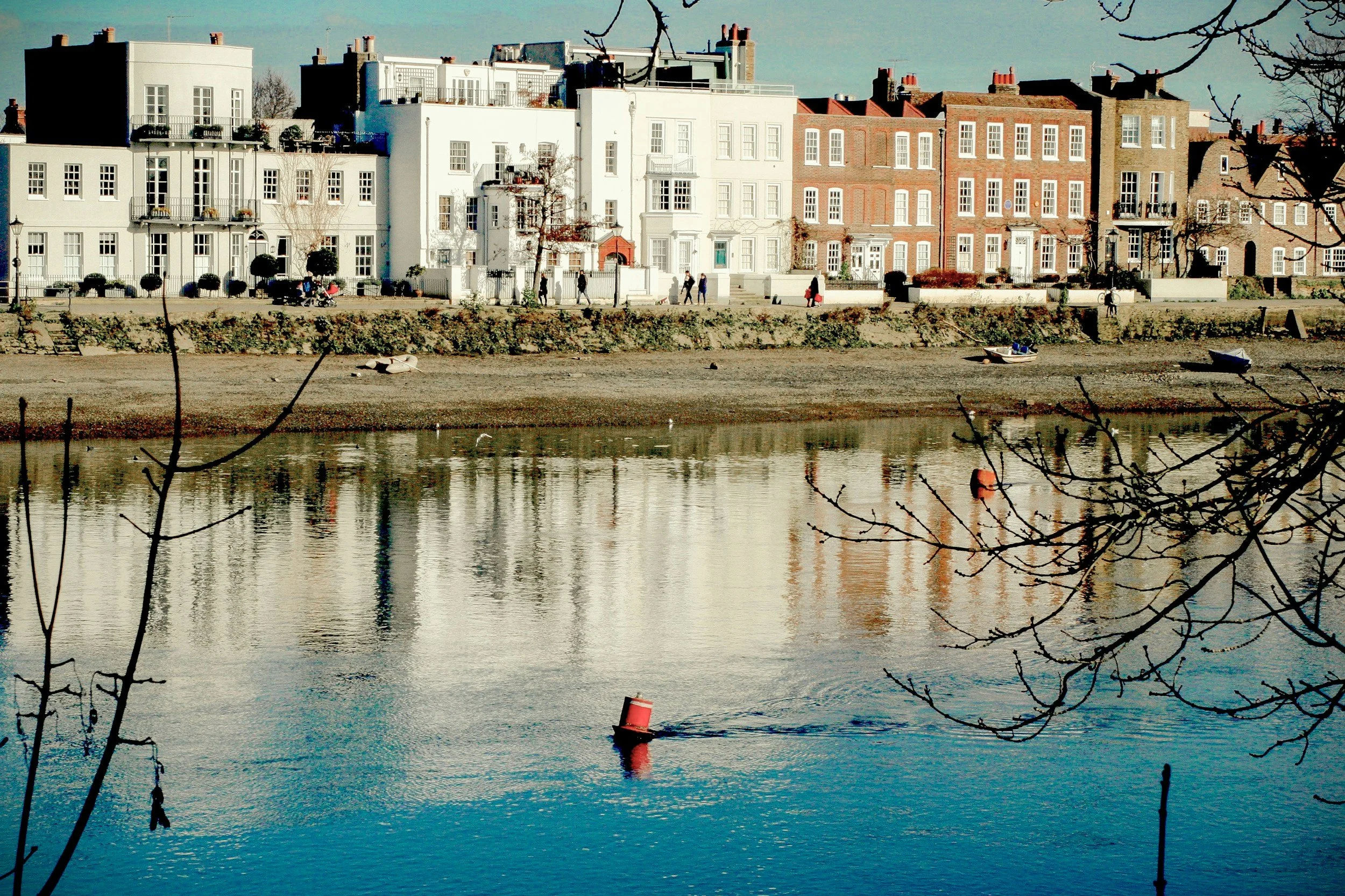 A row of colorful residential buildings along a riverside with boats and people strolling, framed by tree branches.