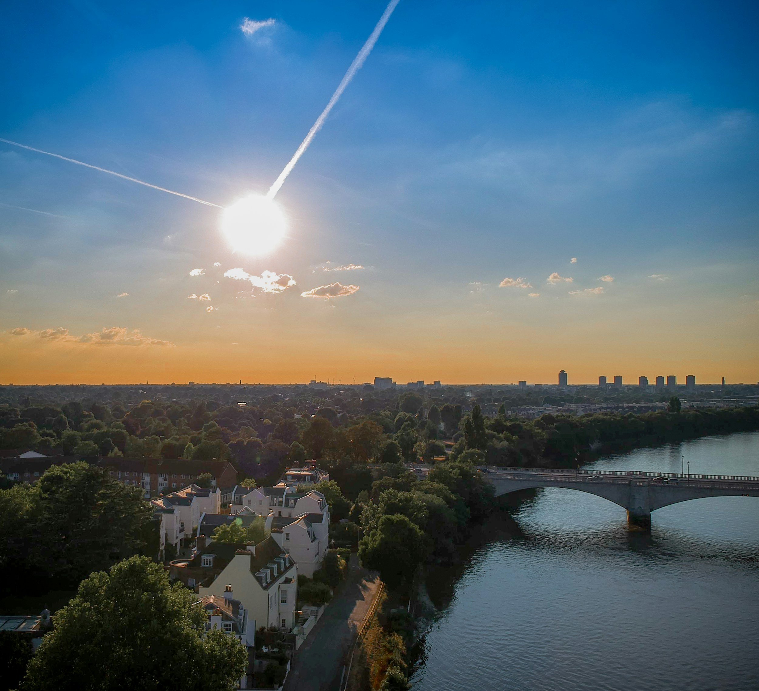 Sunlit view of a river with a bridge, surrounded by trees and residential buildings under a clear blue sky with contrails.