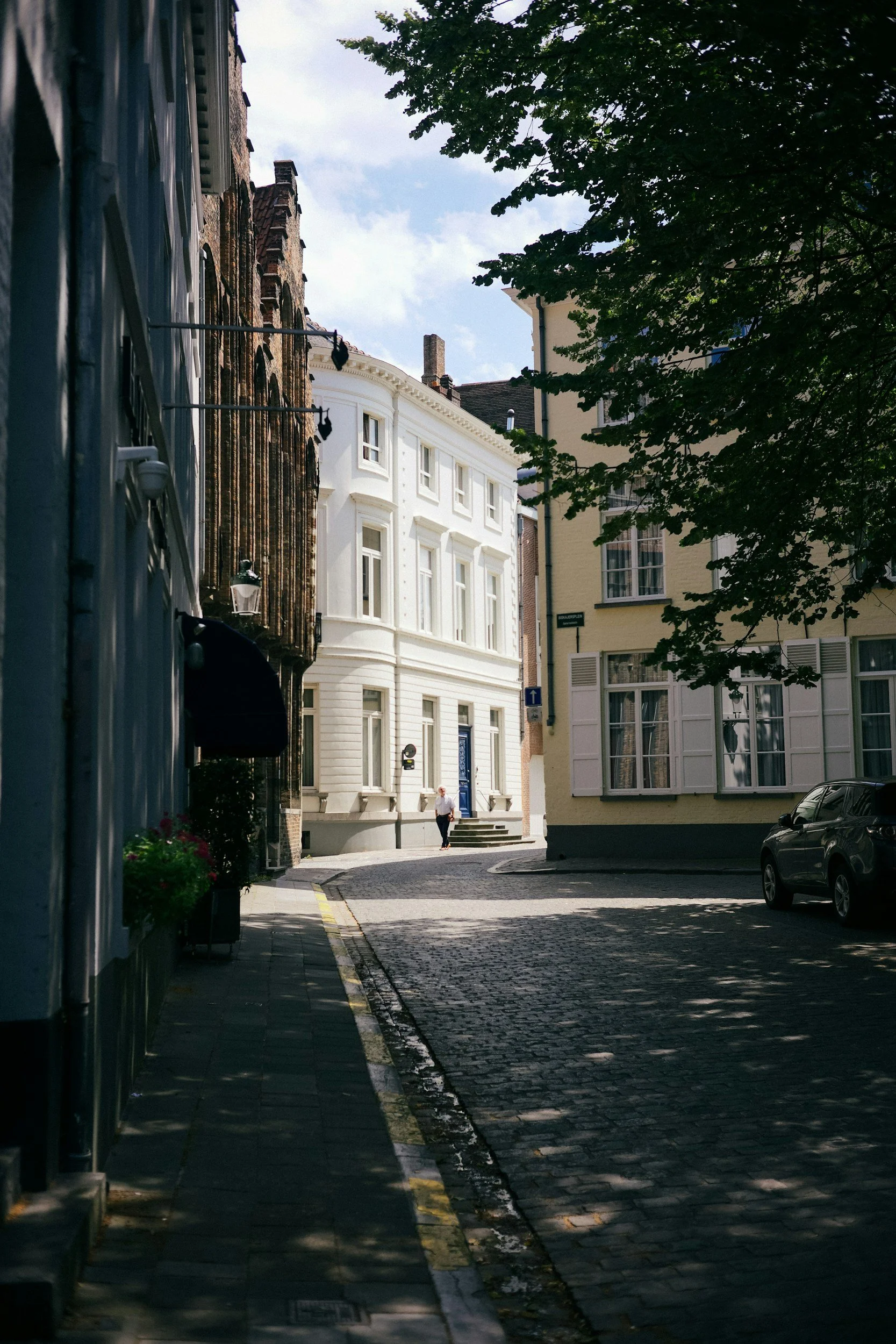 A cobblestone street in a European city with pastel-colored buildings, a person walking in the distance, and shadows of trees on the road.