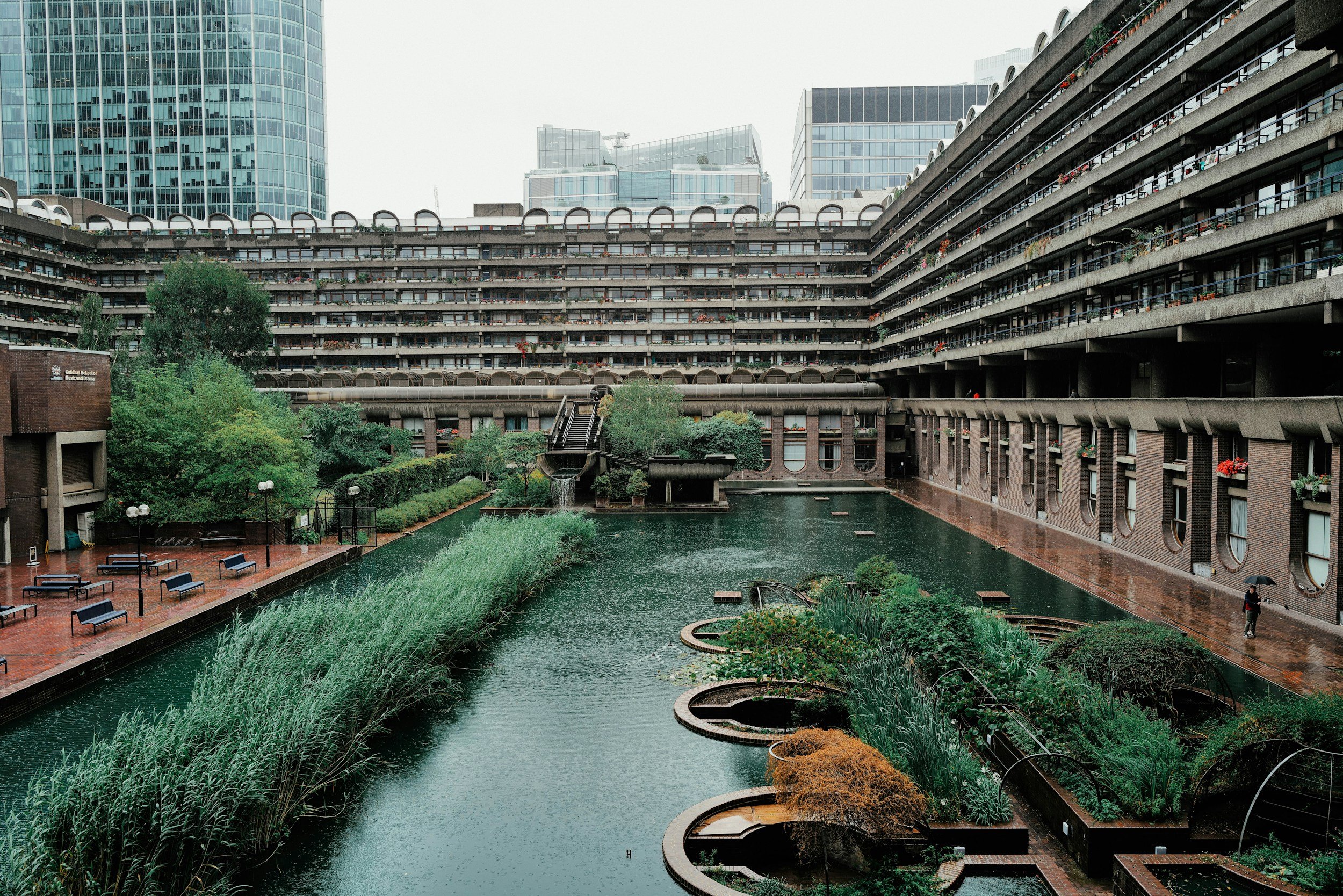 View of a courtyard with a water feature surrounded by tall modern buildings, with few people walking in the rainy weather.