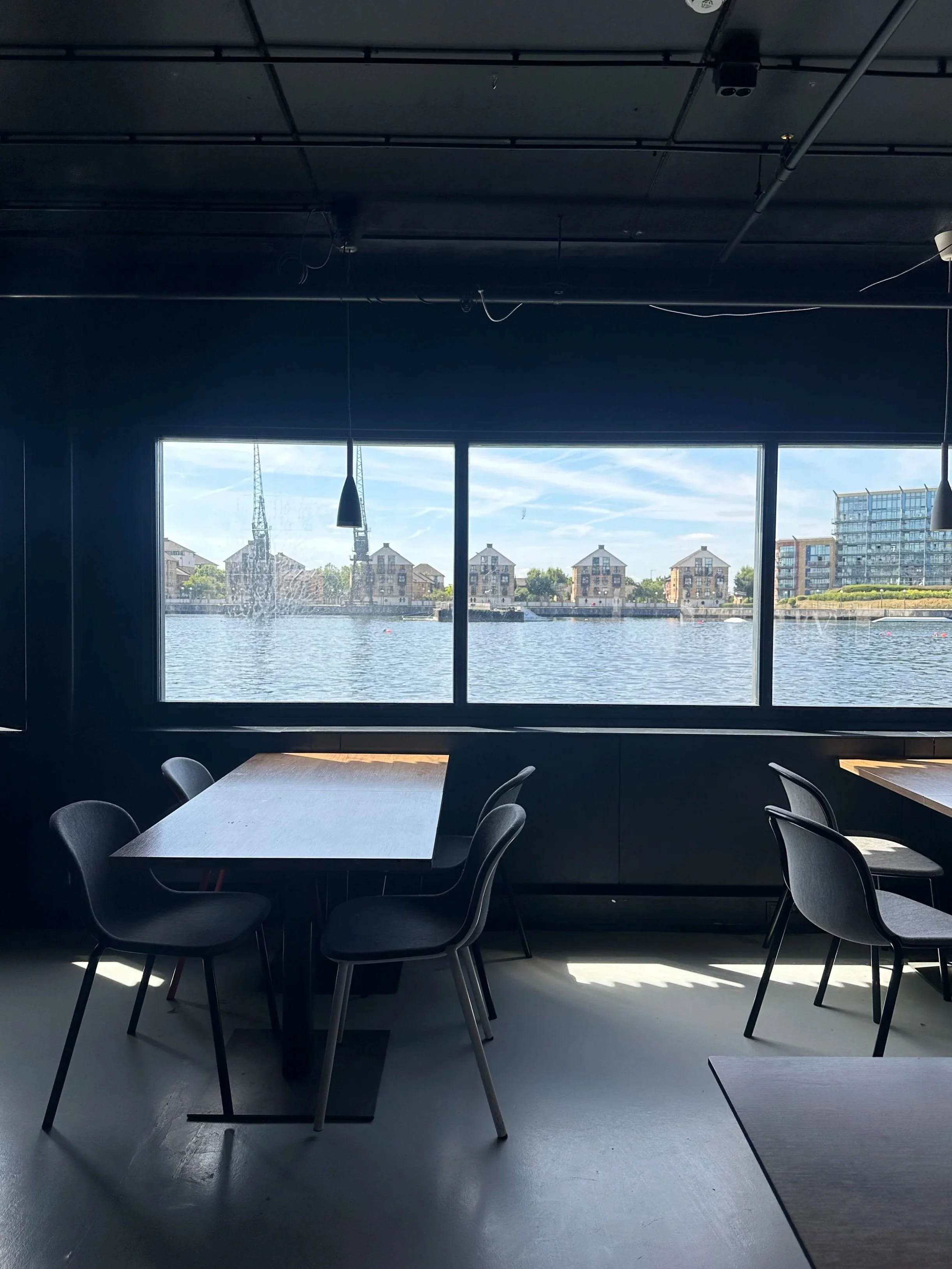 Interior of a modern restaurant or cafe near a waterfront with large window views of water and residential buildings across the river.