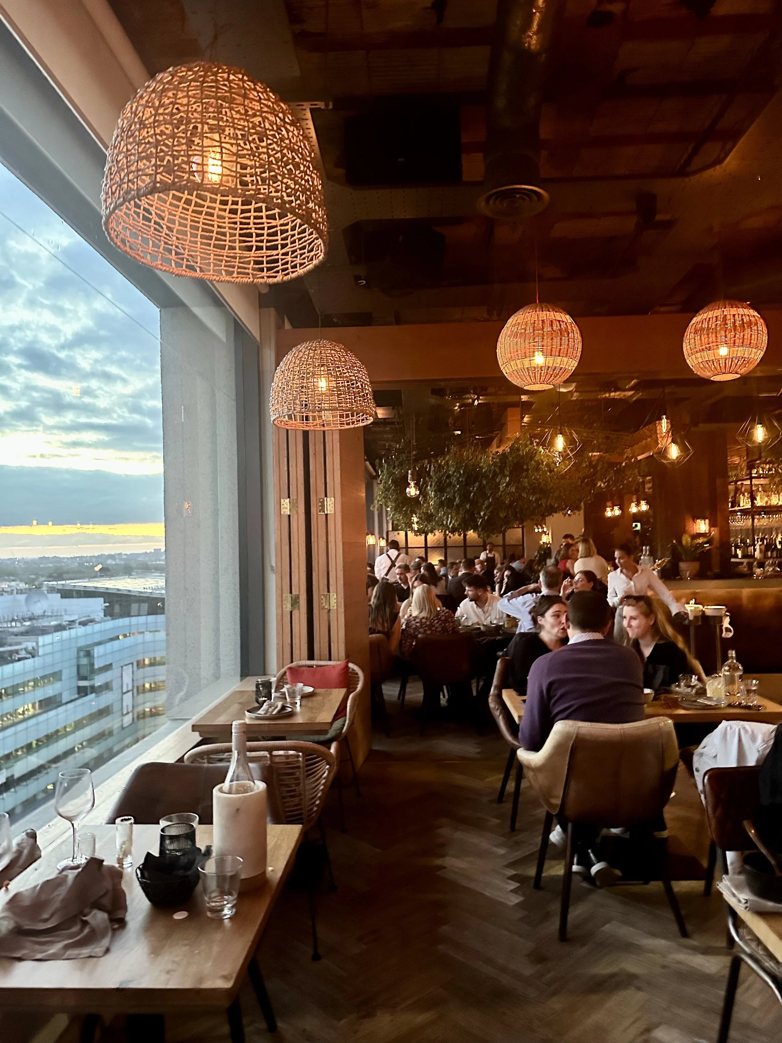 Interior of a restaurant with hanging woven pendant lights, large windows showing a cityscape and evening sky, and patrons dining at tables.