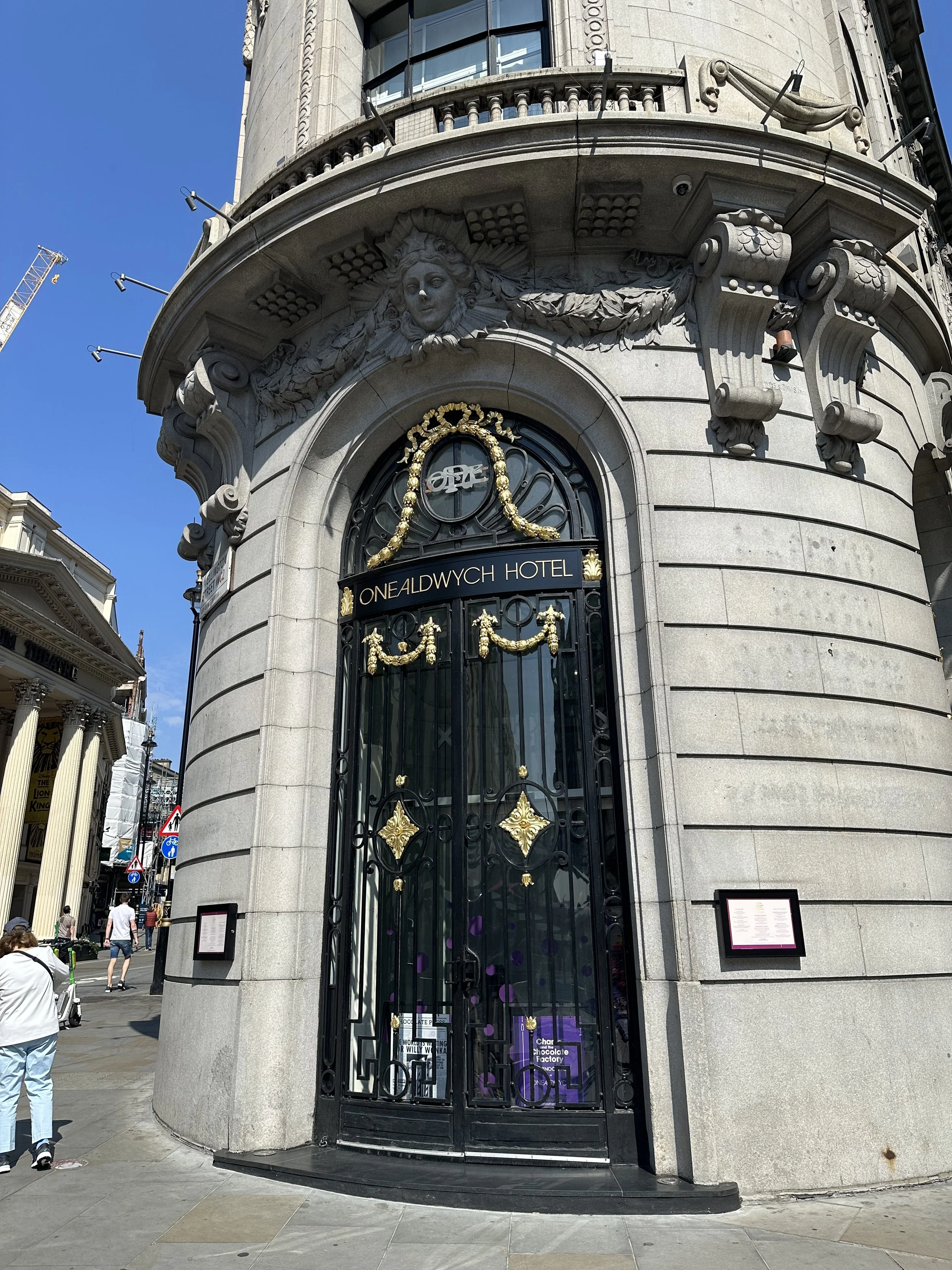 The entrance to the One Aldwych Hotel, a historic building with ornate stonework and decorative gold accents on the black iron gate, located on a busy city street with pedestrians and nearby structures.