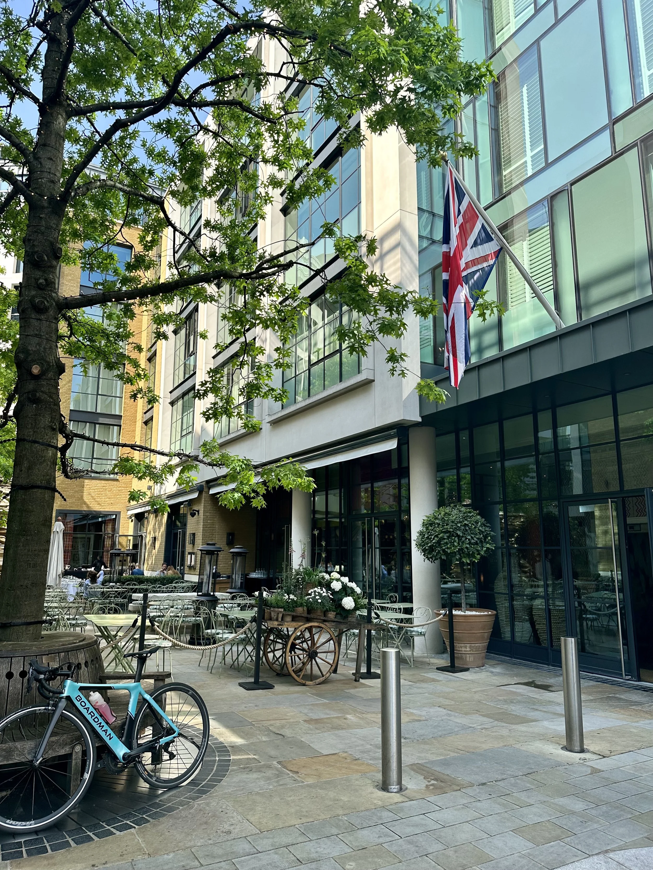Outdoor patio of a restaurant or cafe with tables and chairs, a potted tree, a bicycle labeled 'Boardman,' a wagon with white flowers, and an American flag flying outside a modern building with large glass windows, trees, and other buildings in the background.
