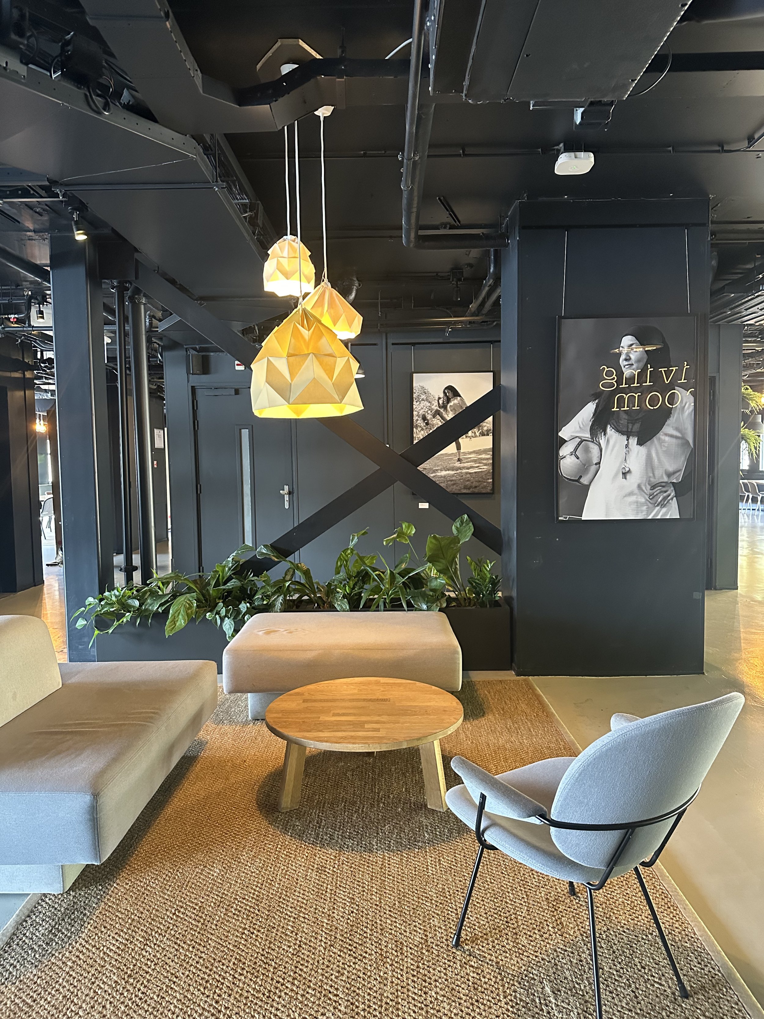 Modern hotel lobby with beige sofa, white armchair, round wooden coffee table, green plants, black walls, black and white artwork, and hanging geometric yellow pendant lights.
