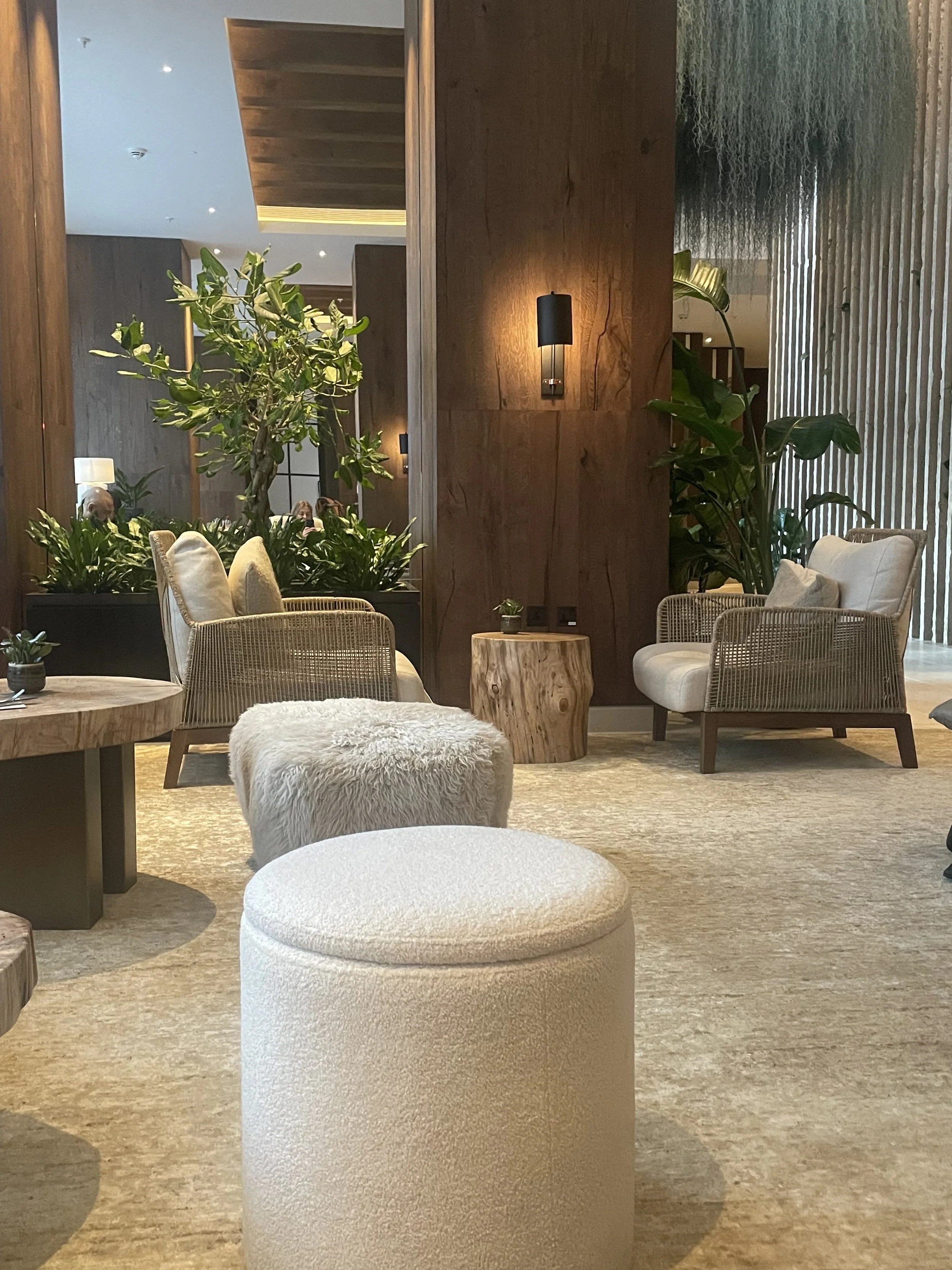 A cozy hotel lobby with beige and cream upholstered furniture, wooden accents, indoor plants, and warm lighting.
