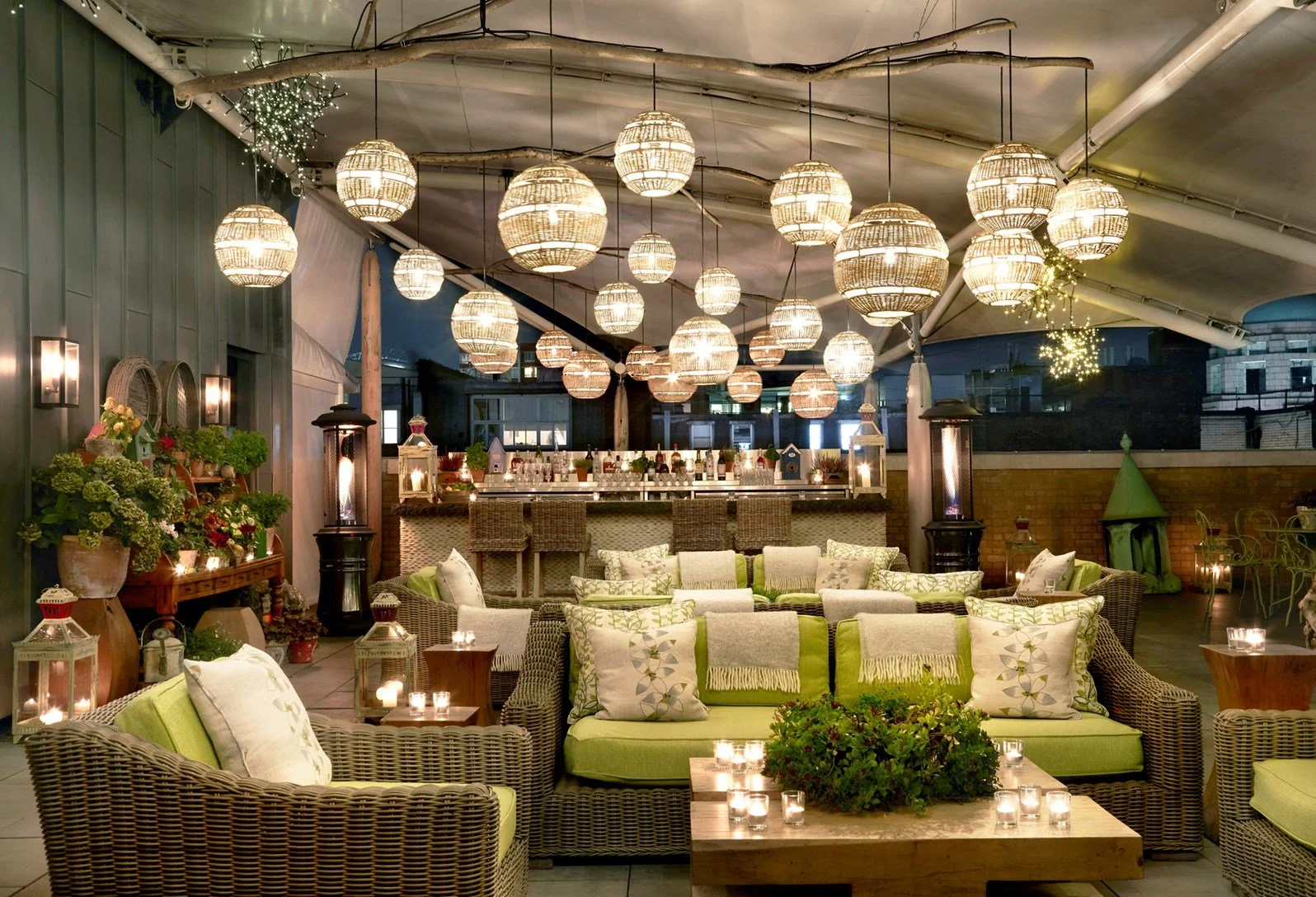 An evening outdoor lounge area decorated with hanging string lights, lanterns, and candles on tables. Wicker sofas with green and white cushions, potted plants, and a bar in the background.