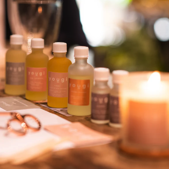 A row of small bottles labeled 'You & i' on a table, with a lit candle in the foreground and blurred background of outdoor setting.