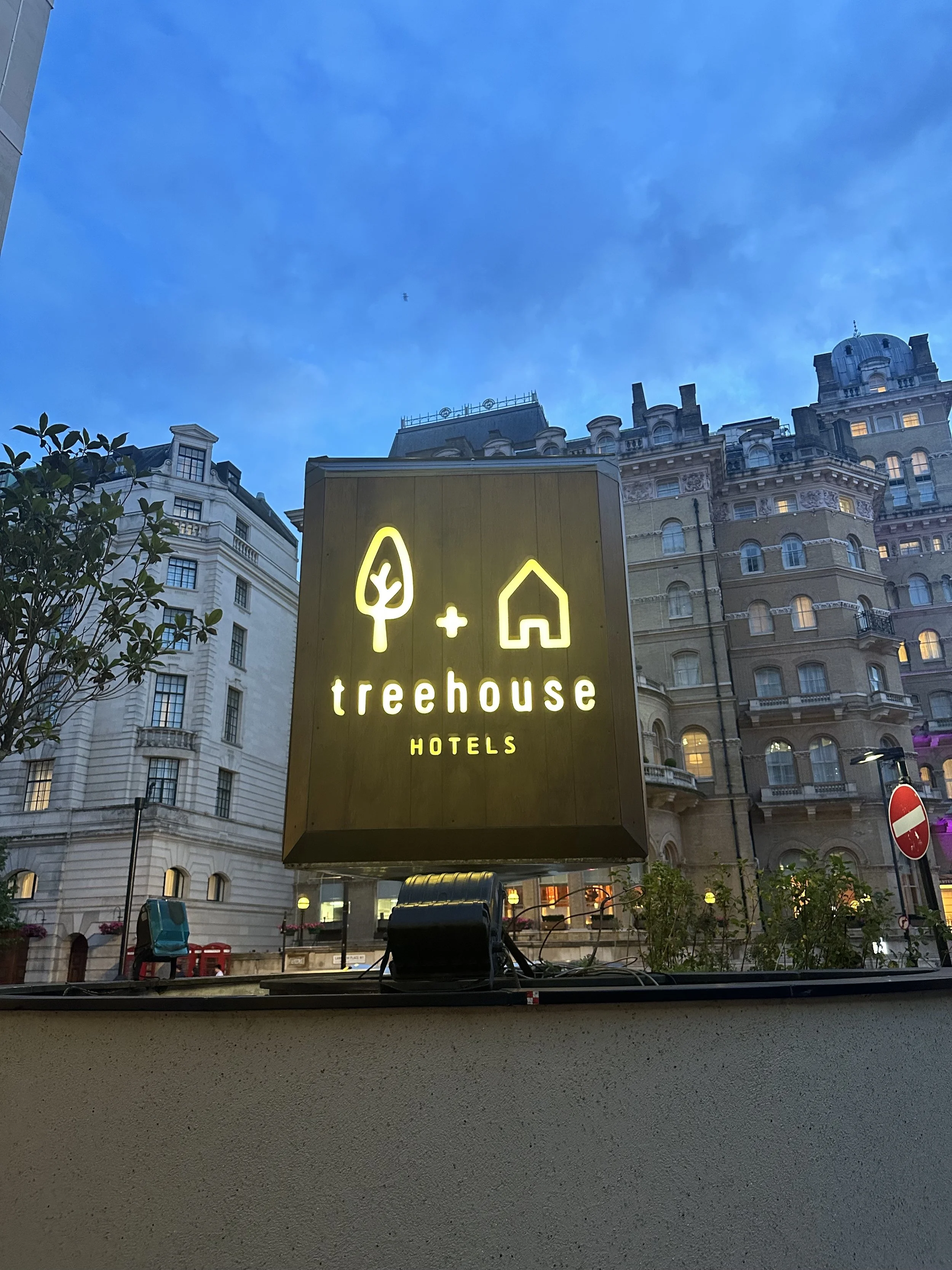 A sign for Treehouse Hotels featuring a tree and house icon, with surrounding buildings and a blue sky.