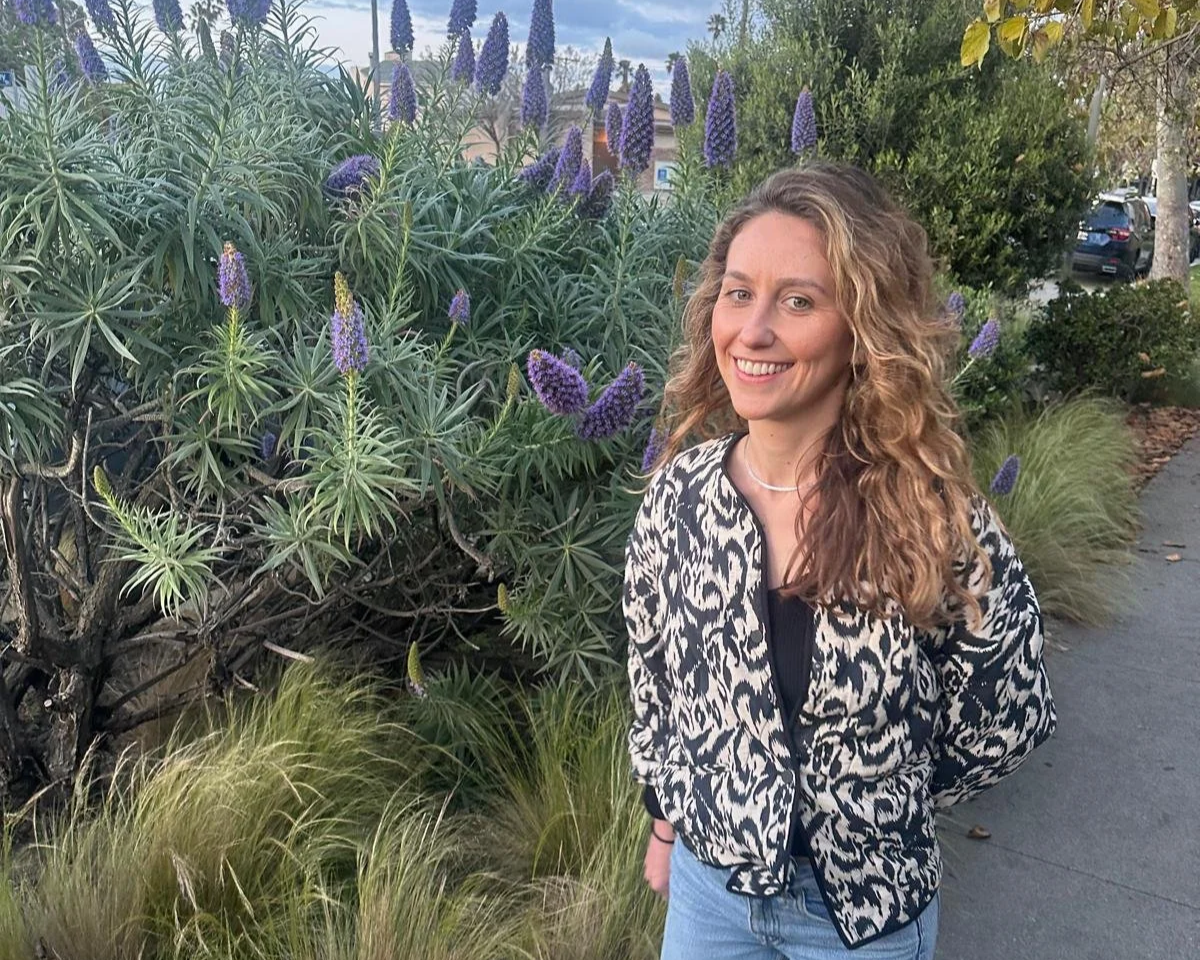 A young woman with curly blonde hair smiling outdoors beside purple flowering plants and greenery.