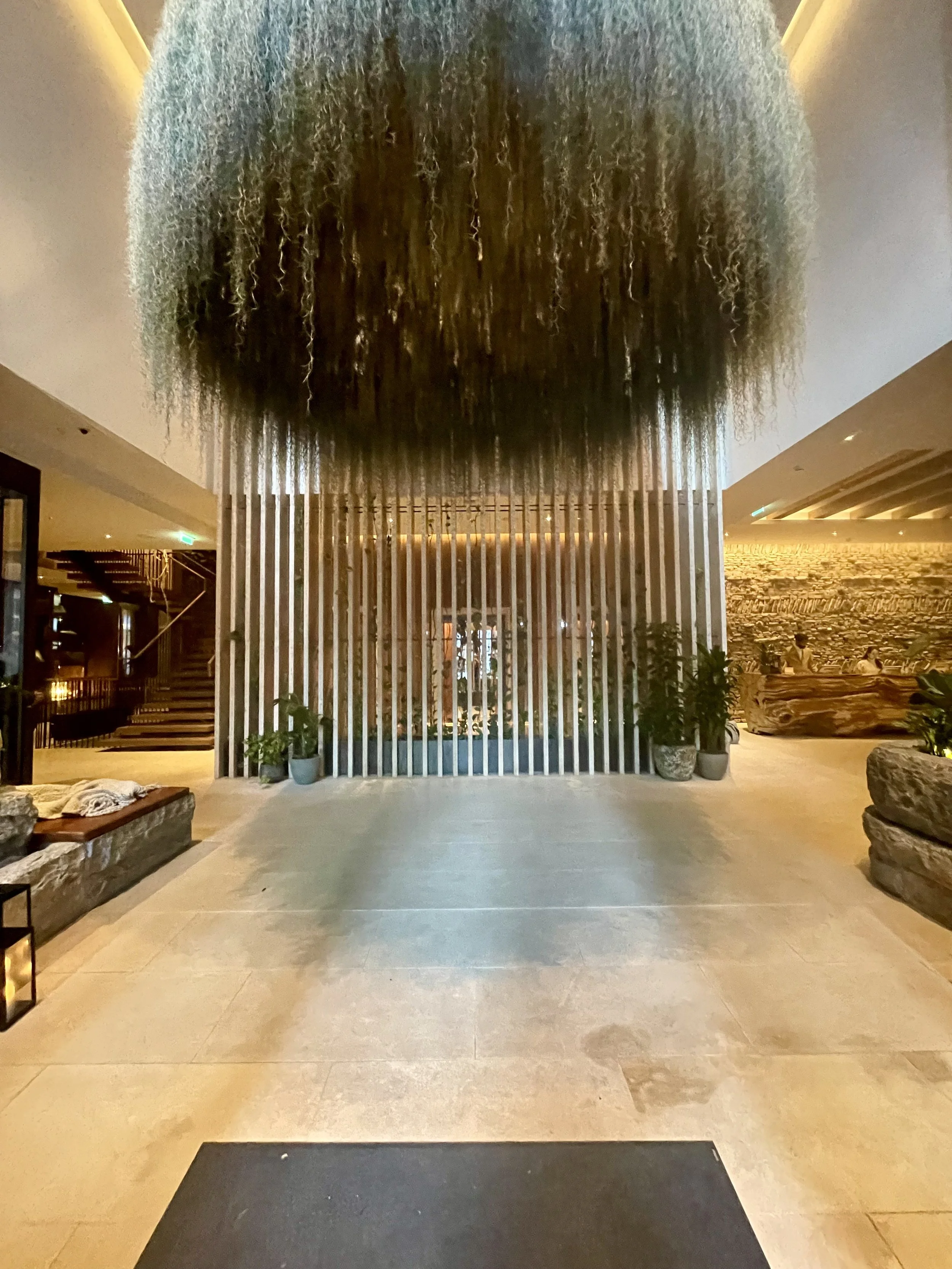 Interior view of a modern hotel lobby with a large chandelier hanging from the ceiling, potted plants, and a stone seating area.