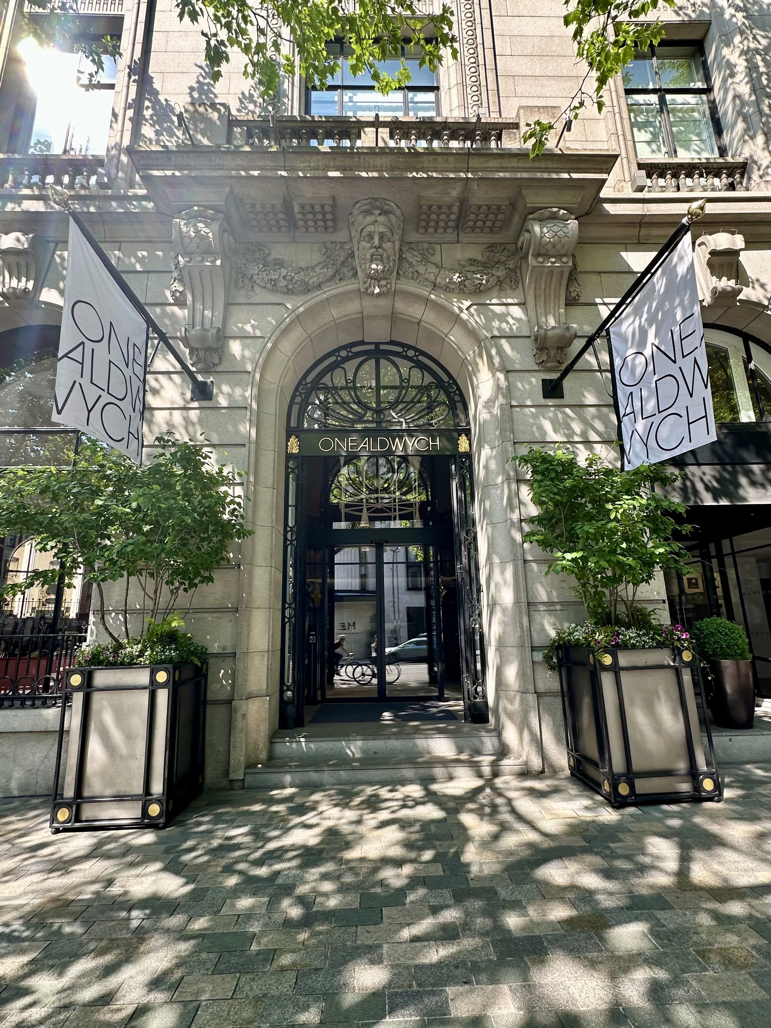 The entrance to a building with the name 'One Aldwych'. The entrance is framed with decorative stonework including a sculpted face and floral motifs. There are two potted plants on either side of the stairs leading to the door. Two flags with the words 'One Aldwych' are hanging on poles attached to the building.