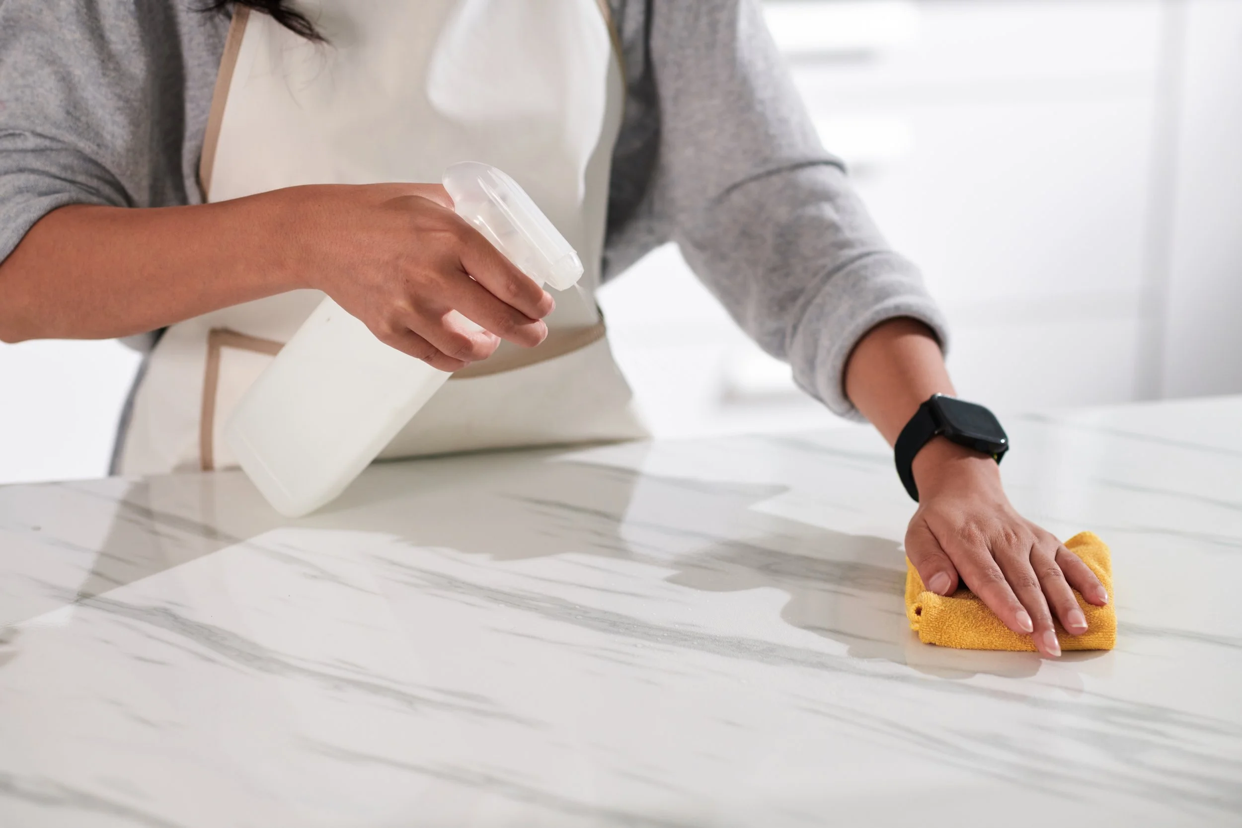 Woman in an apron spraying cleaner on a countertop and wiping it down with a microfiber towel