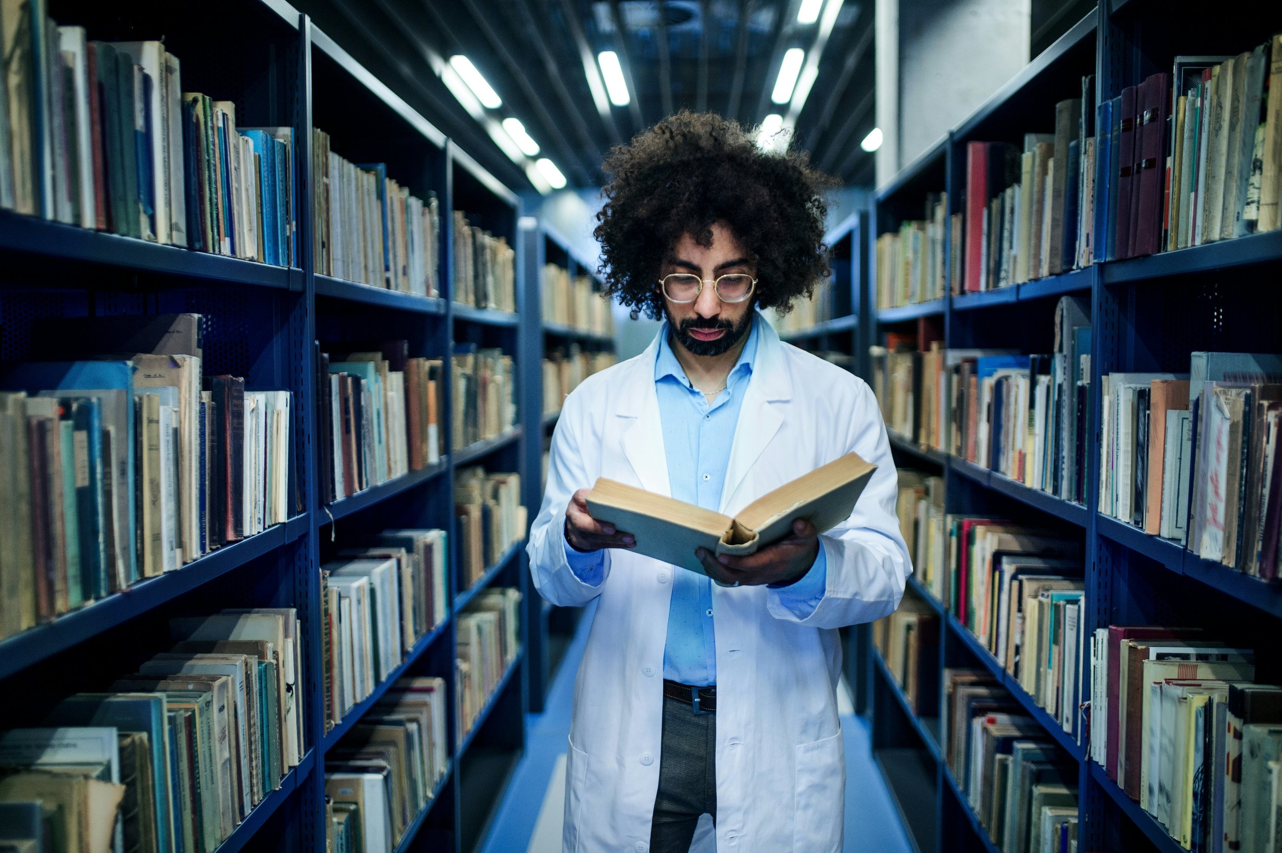 Man in a white laboratory coat reading a textbook amidst the stacks in a library