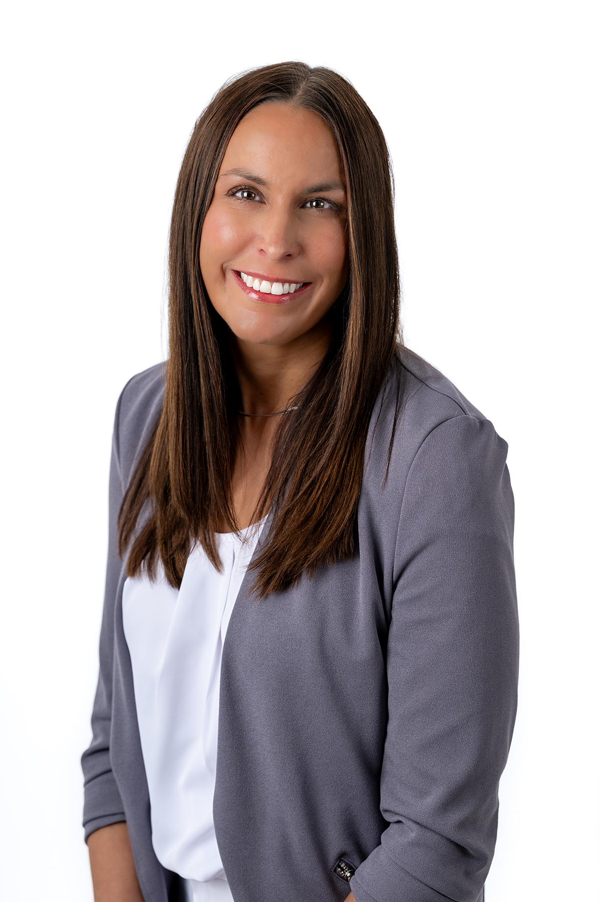 Portrait of a woman with long brown hair smiling, wearing a gray blazer over a white top, against a white background.
