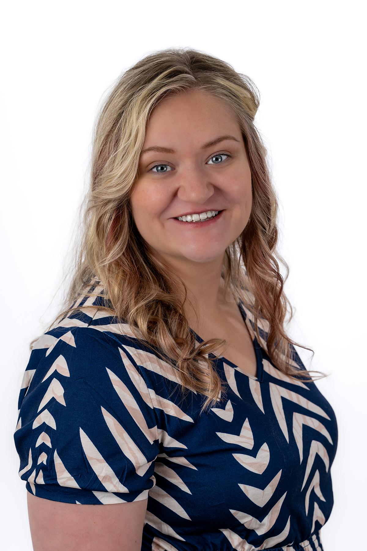 Headshot of a smiling woman with blonde curly hair, wearing a blue and beige patterned dress, against a plain white background.