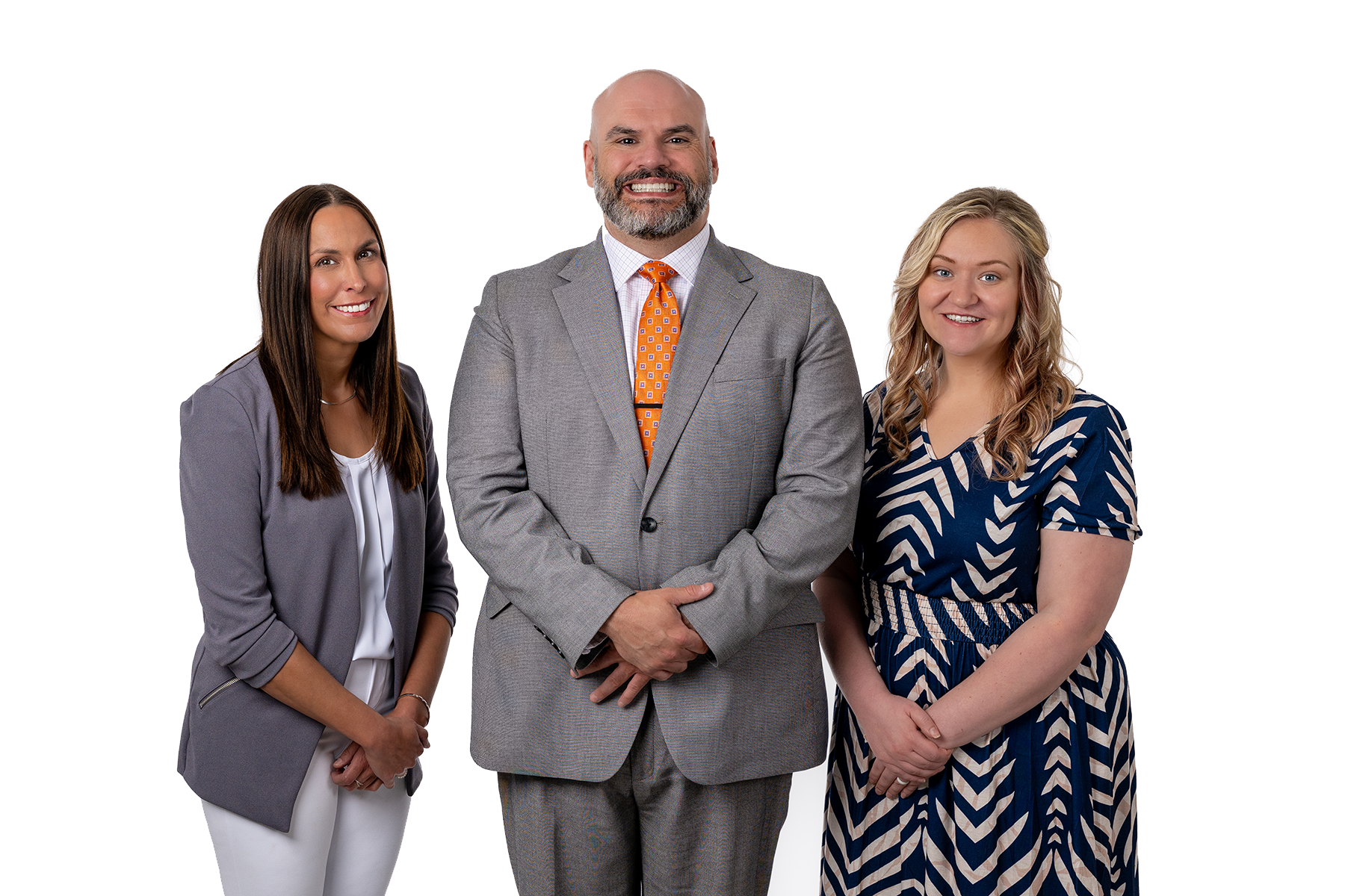 Three professionally dressed individuals posing for a photo against a plain white background: a woman in a gray blazer and white outfit on the left, a man in a gray suit and orange tie in the middle, and a woman in a blue and cream patterned dress on the right.