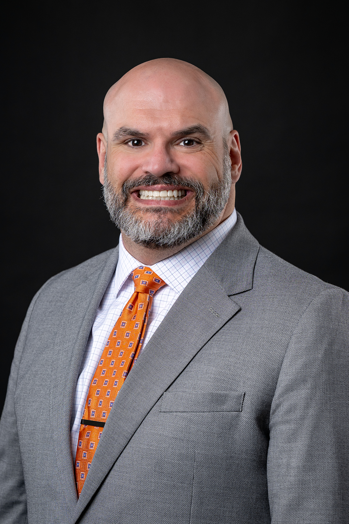 A professional man with a bald head, gray beard, and mustache, wearing a gray suit, white checkered shirt, and orange patterned tie, smiling against a dark background.