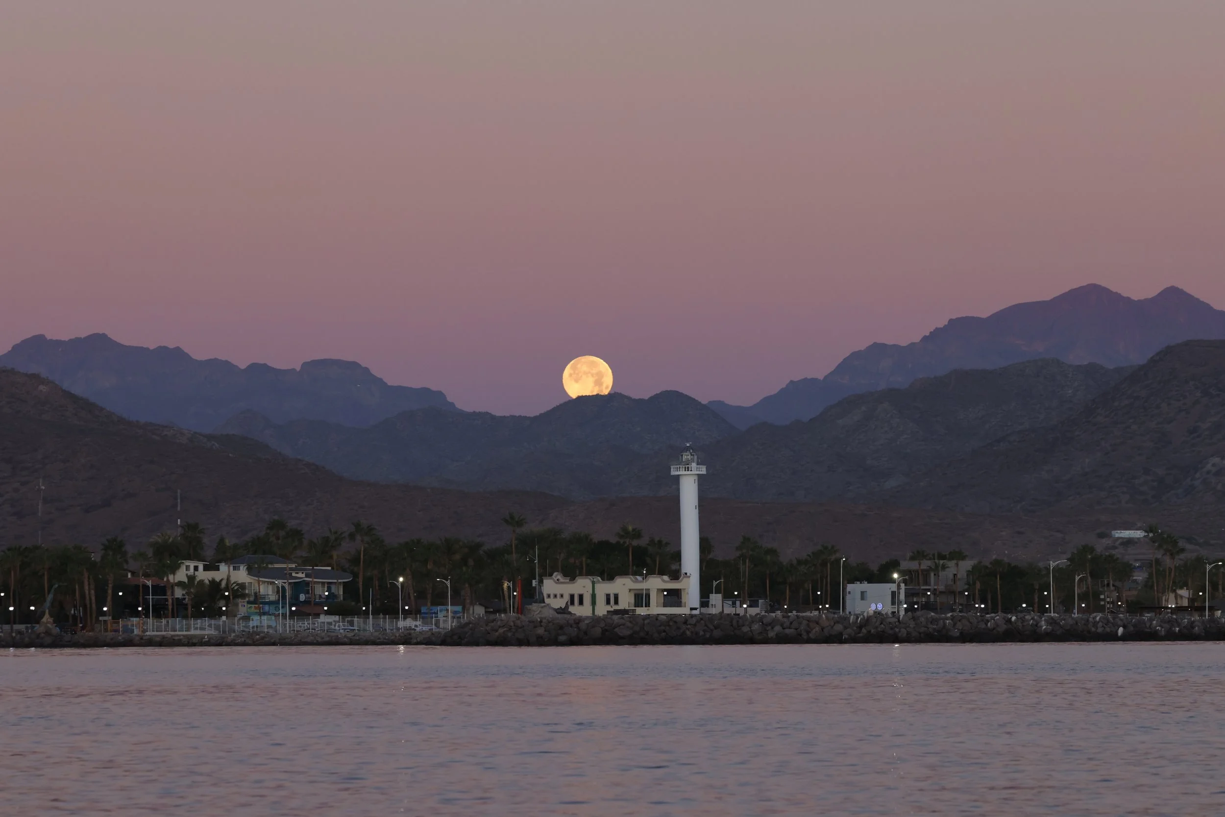 Loreto lighthouse on a waterfront with mountains in the background under a pink sky and full moon, in Loreto Bay National Park, Baja California Sur, Mexico during a whale watching tour