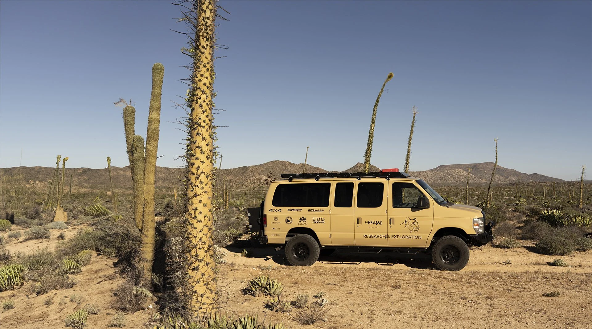 A beige off-road vehicle parked in a desert landscape with cactus plants and mountains in the background.