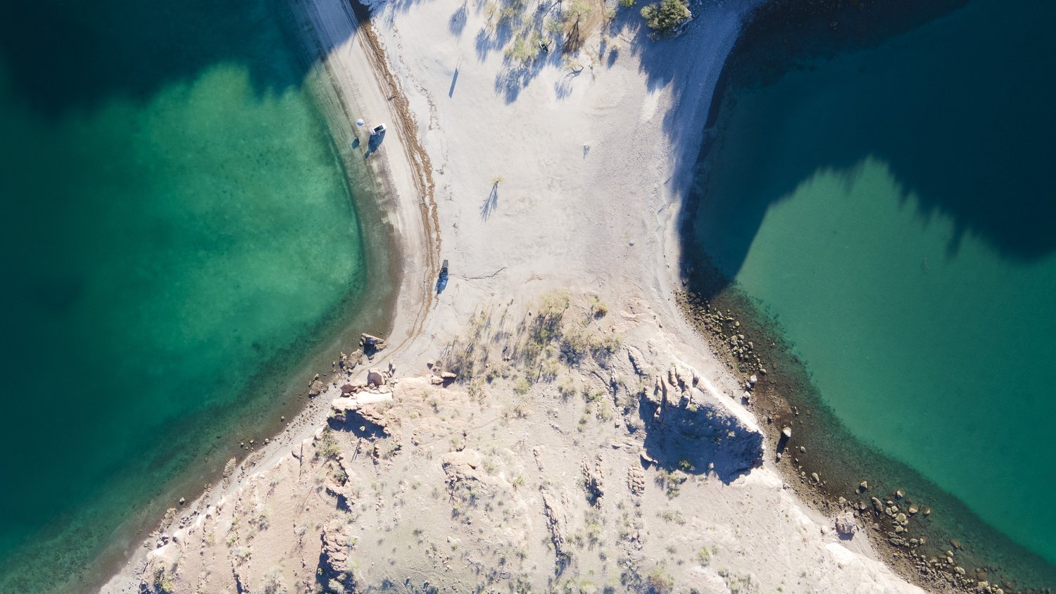 Aerial view of a narrow strip of land between two bodies of water, one greenish and one dark blue, with boats on the shore and shadows cast by trees and cliffs.